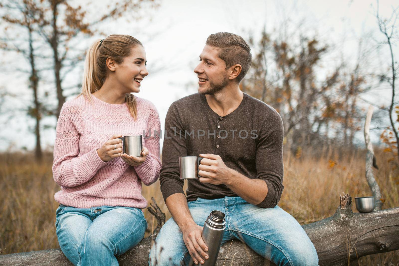 Young Couple Communicate Happily With Each Other Drinking Hot Drink In Nature, Cheerful Woman And Man Have Rest Sitting On Log And Holding Cups Of Hot Drink, Coffee Break Outdoors Concept
