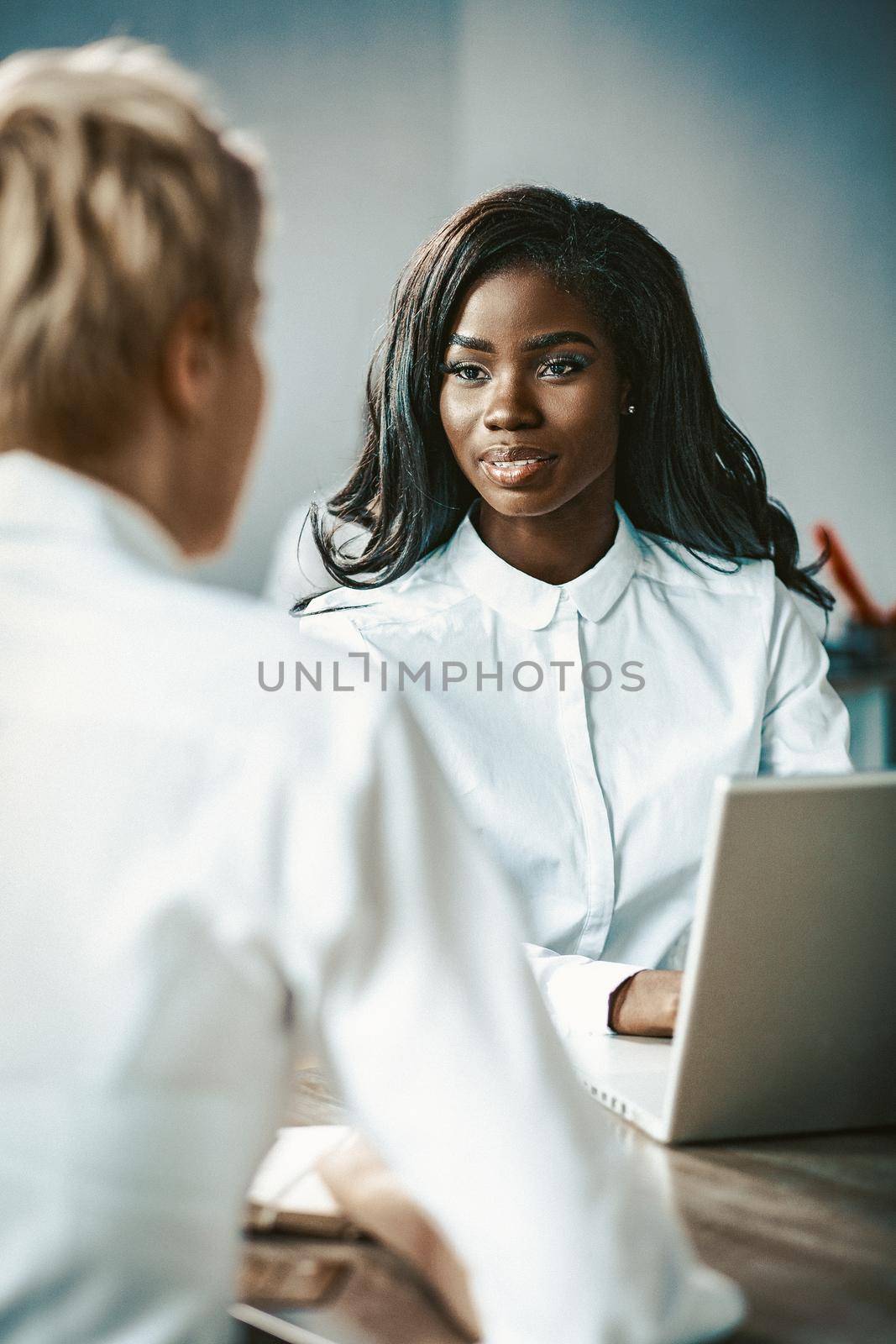 Two Business Women Talking Each Other, Caucasian And African Ladies Talks Sitting At Table, Focus On Face Of Charming African Woman Chatting With Blonde Sitting Back In Foreground, Toned Image