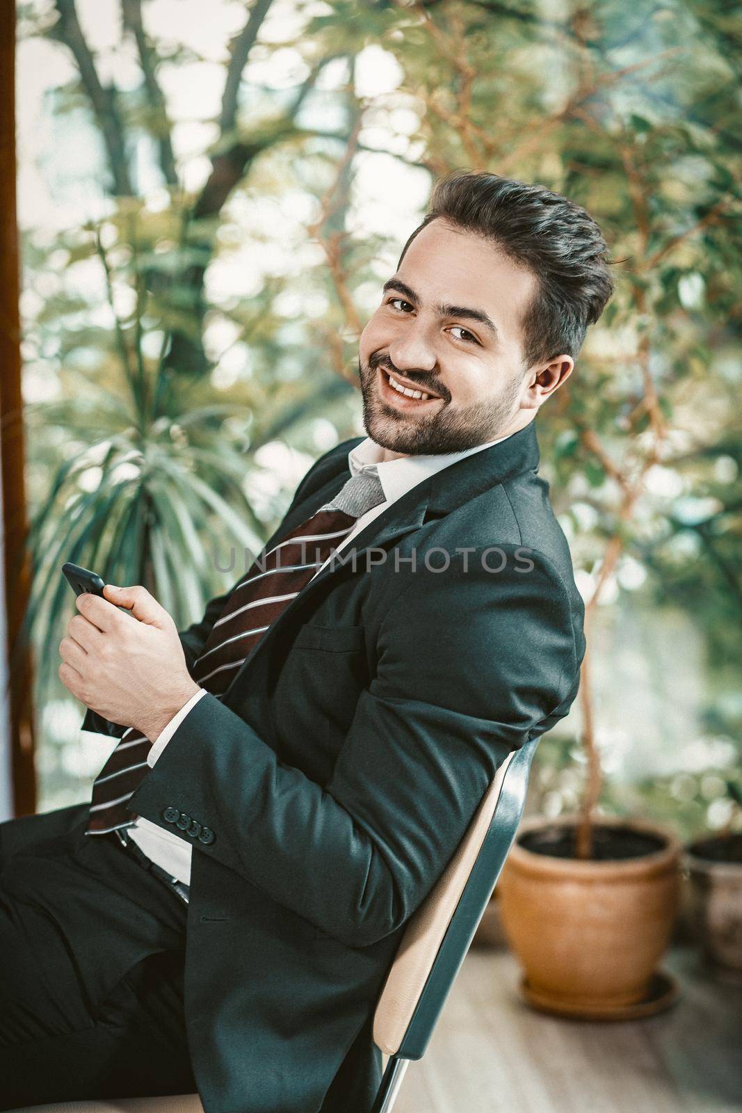 Happy businessman holds Phone Looking At Camera , Portrait Of Well-dressed Caucasian Man Typing On Smart Phone During Coffee Break While Sitting At Office Chair On Floral Background, Toned Image