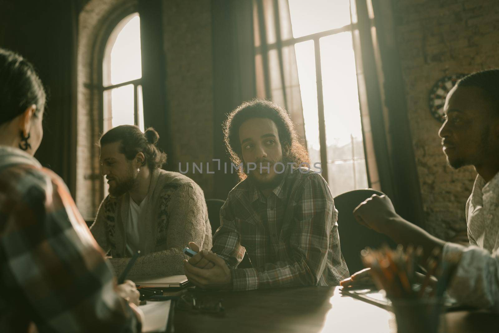 Team Of Young Freelancers Working Together In Workspace, Focus On Arab Guy Sitting In Back Sunlight, Diverse Team Communicates At Table In Stylish Interior With High Arched Windows