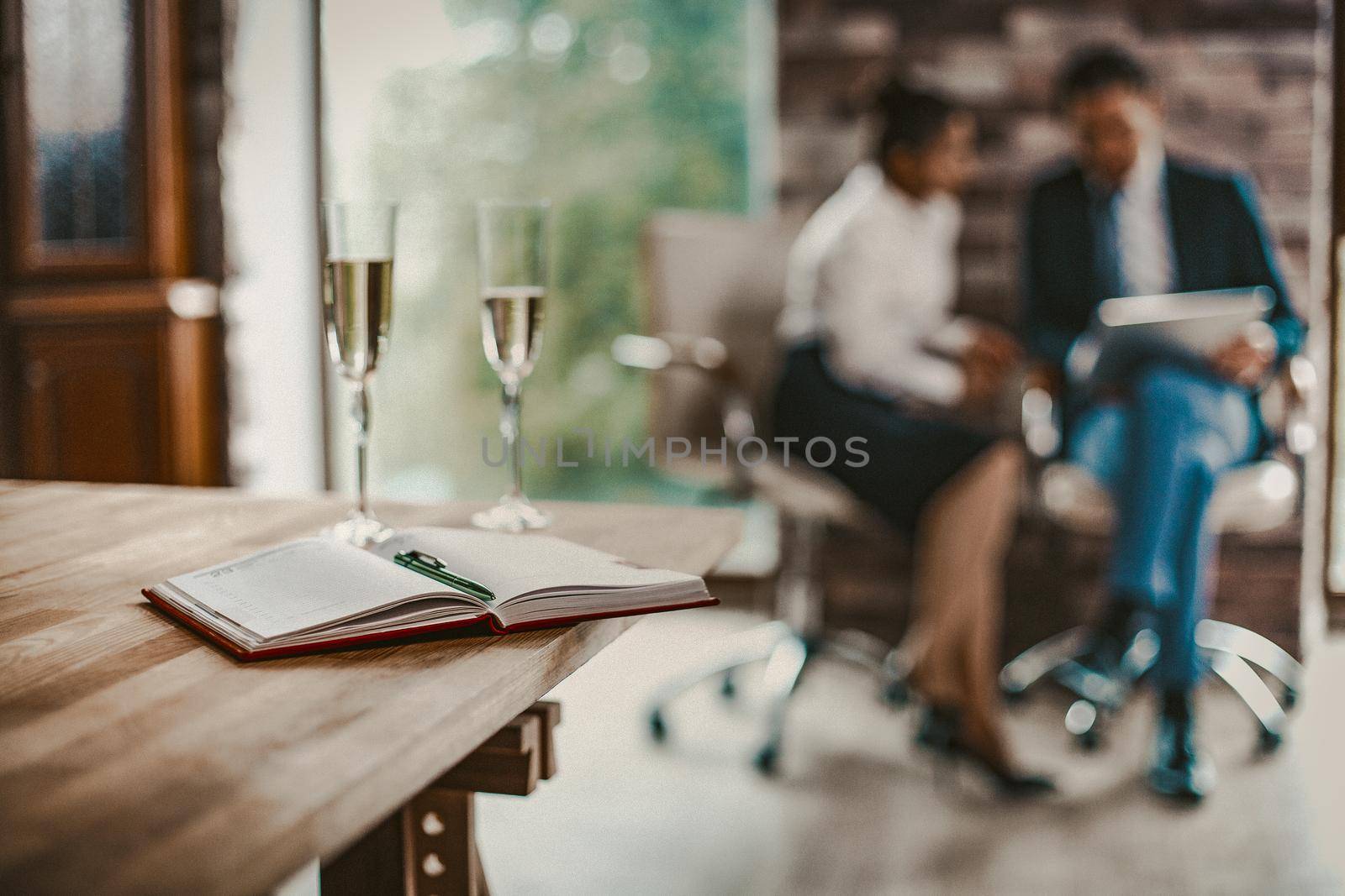 Selective Focus On Opened Notepad With Pen Laying On Wooden Desk In Foreground, Wine Glasses Of Champagne Standing On Table, Business Man And Woman Have Conversation On Blurred Background, Toned Image