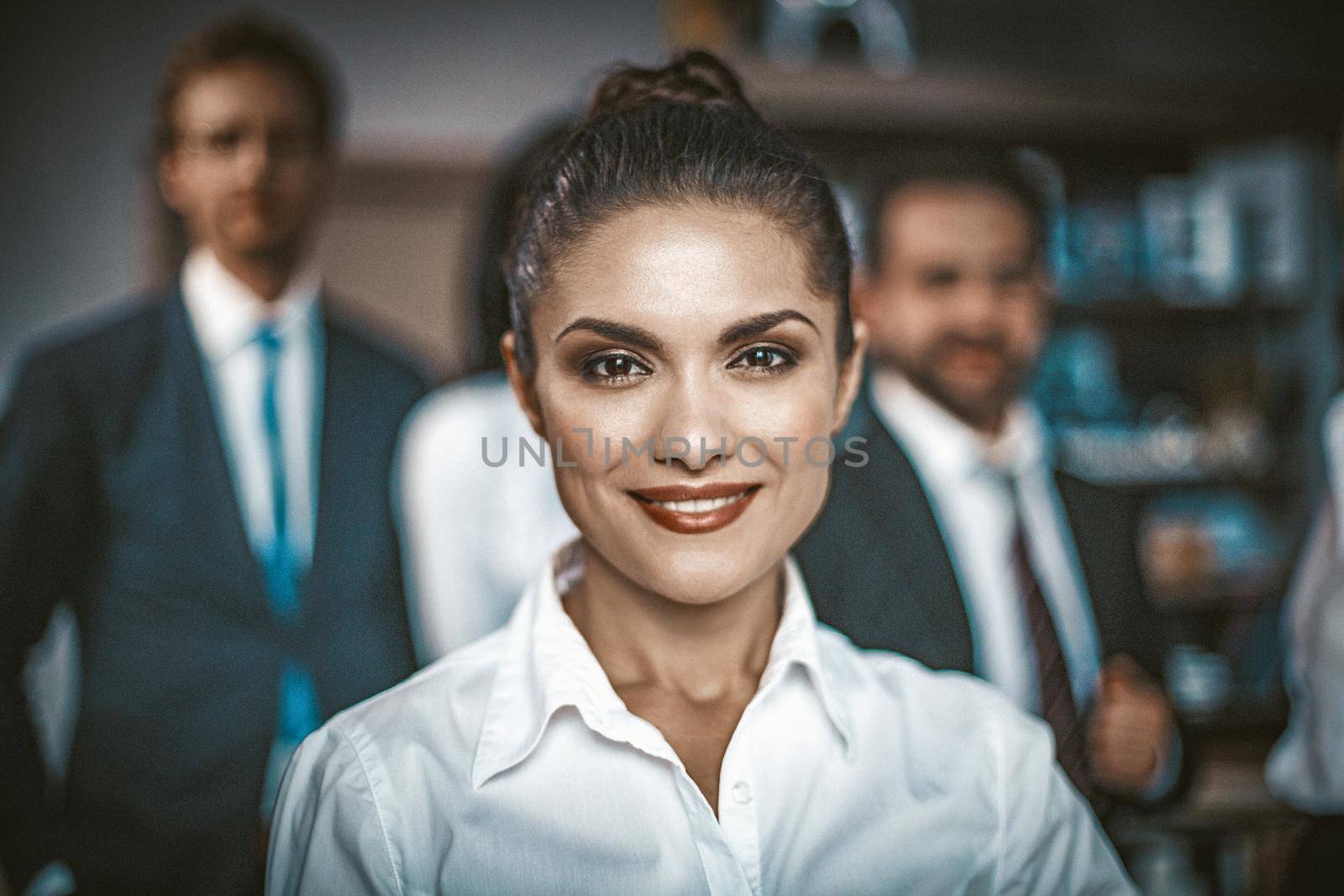 Colleagues In Suits On Background. Young Office Worker Smiling While Looking At Camera, Group Of Her Male Colleague Are On A Blurred Background, Toned Image