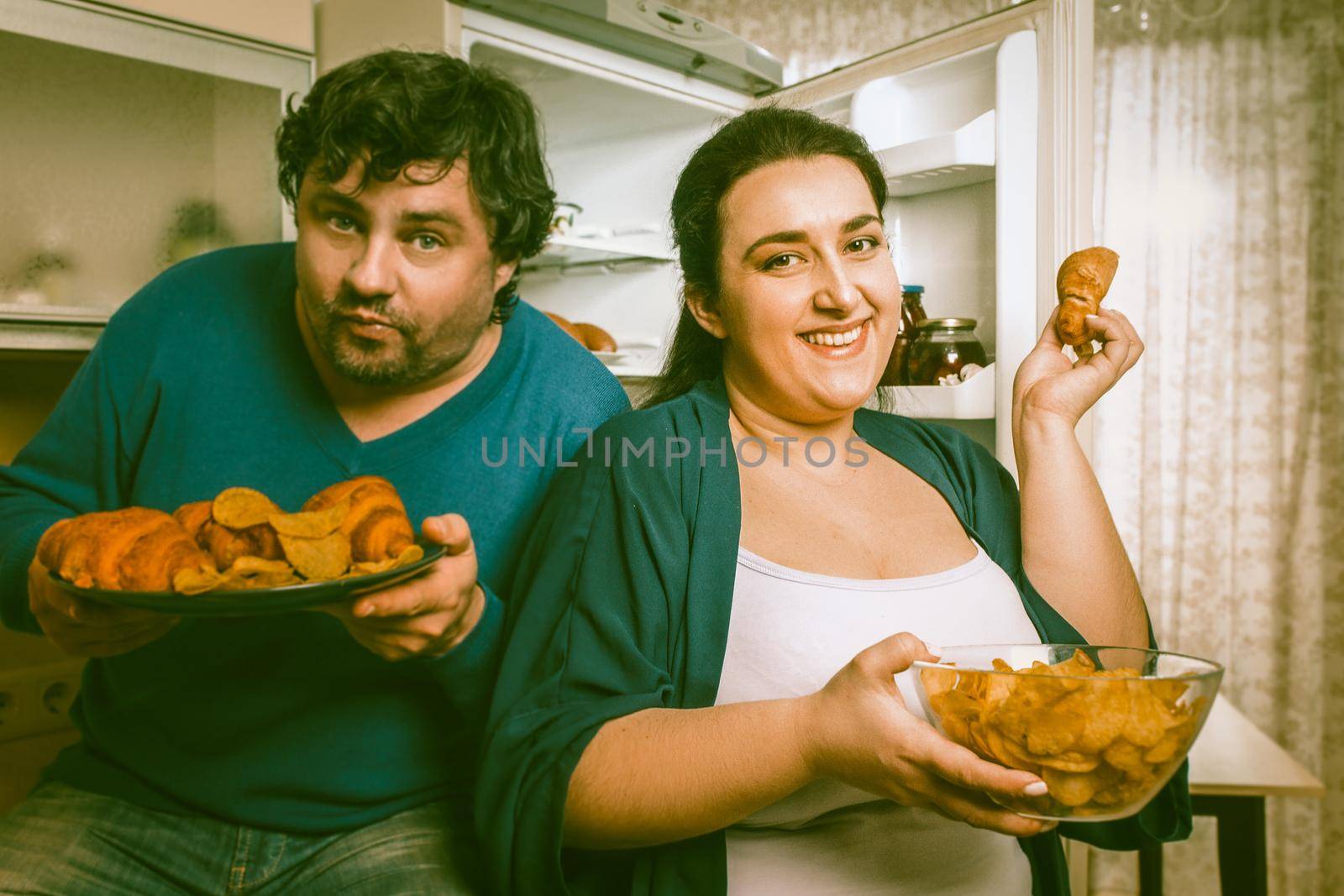 Body Positive Couple Having Fun With Unhealthy Eating In Kitchen, Smiling Man And Woman Standing Near Opened Refrigerator Holding Plate With Croissants And Bowl Of Potato Chips While looking At Camera