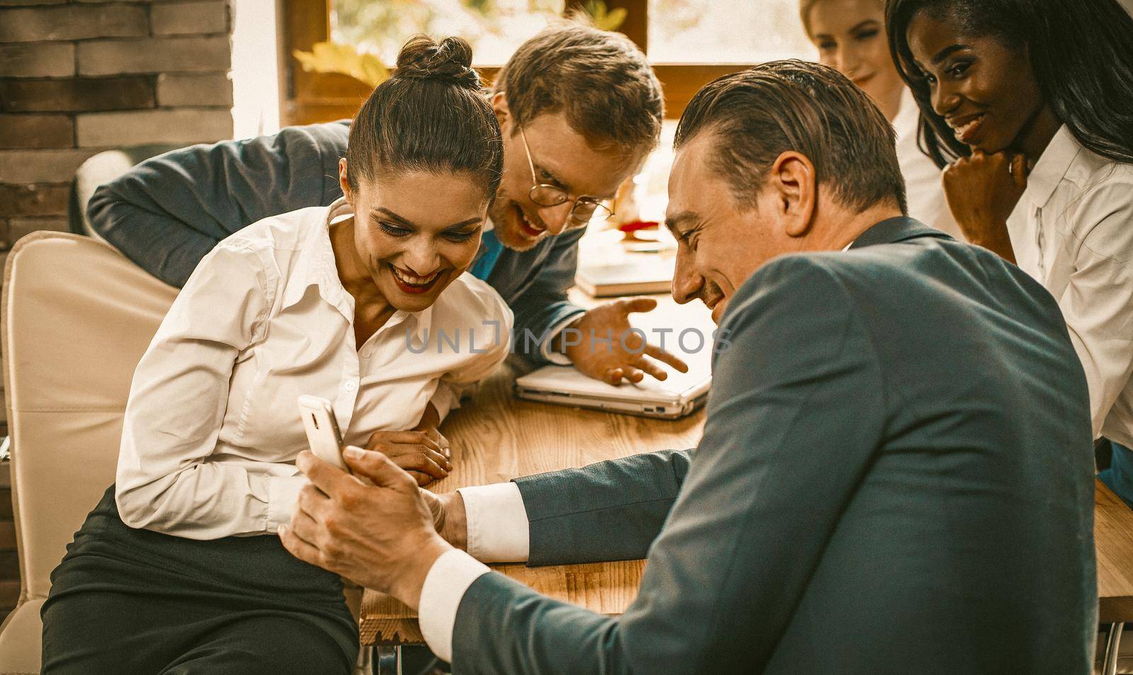 Business Team Of Employees Have Fun Sitting At Table And Using Phone, Charming Young Women In White Shirts And Men Toothy Smiling While Looking At Their Colleague's Phone In Foreground, Toned Image