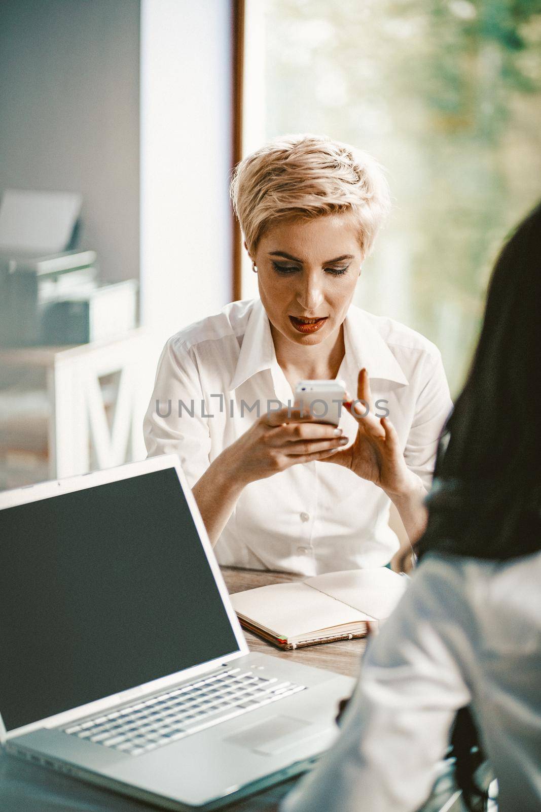 Focused Business Woman Uses Her Phone, Portrait Of Blonde Businesswoman In White Blouse Typing At Mobile Phone During Meeting While Sitting At Table With Opened Notepad And Laptop On It, Toned Image