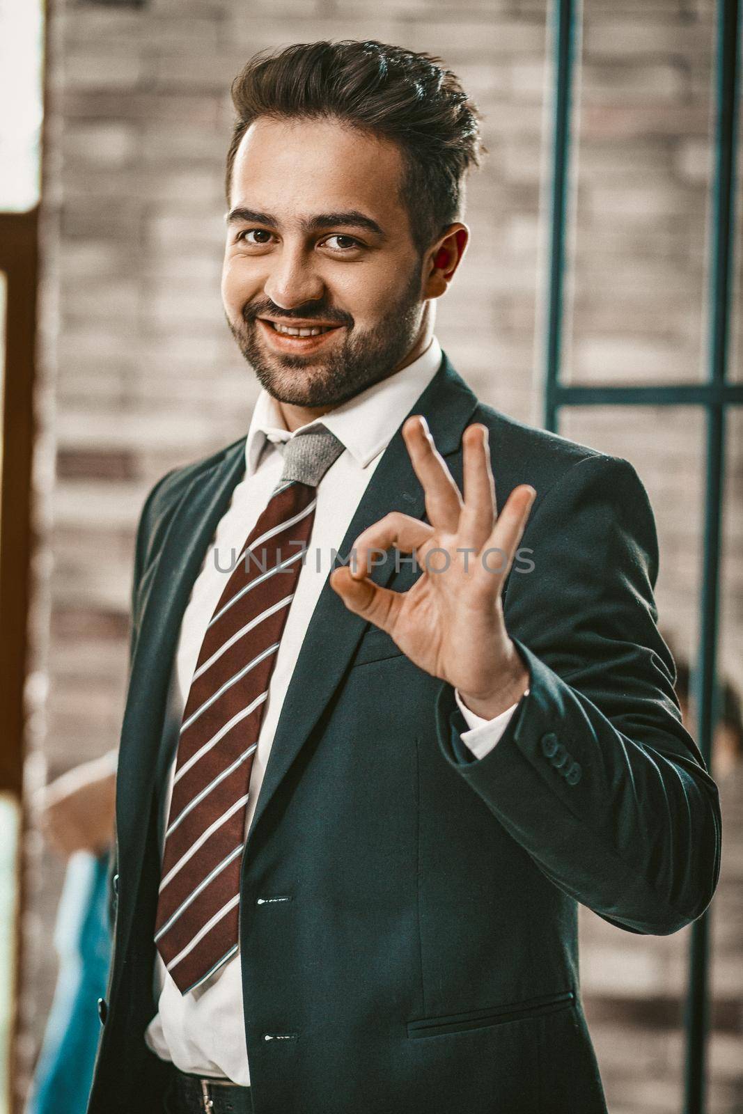 Successful Businessman Shows Okey Gesture, Portrait Of Well-Dressed Bearded Man In Dark Suit Gesturing Ok Sign. Selective Focus On Male Smiling Face, Toned Image