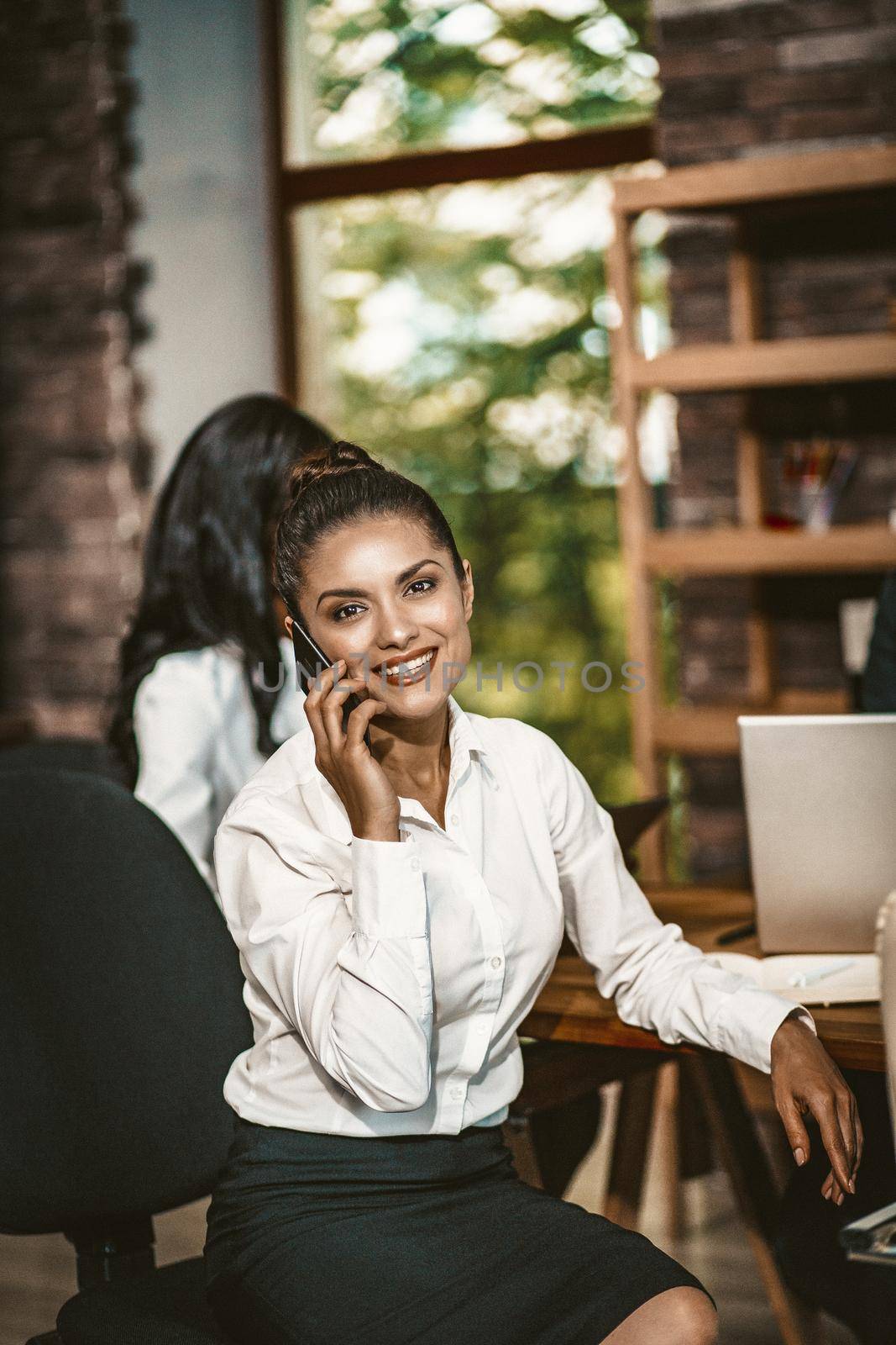 Smiling Business Woman Talks By Mobile Looking At Camera, Charming Brunette In Office Clothes Talking On Phone While Sitting At Office Table During Negotiations, Toned Image