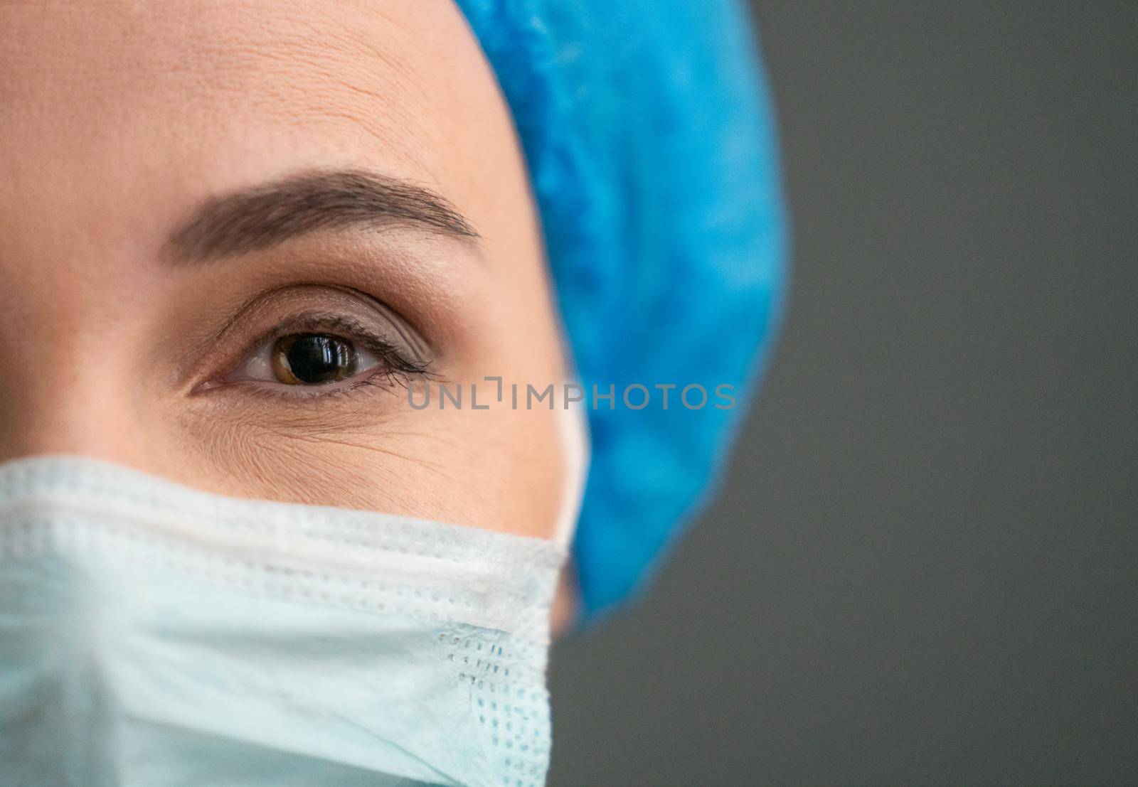 Female medical worker In Protective Mask And Blue Workwear Looking At Camera, Close Up Shot Of Cheerful woman's Eye, Copy Space Are At Right Side On Grey Back