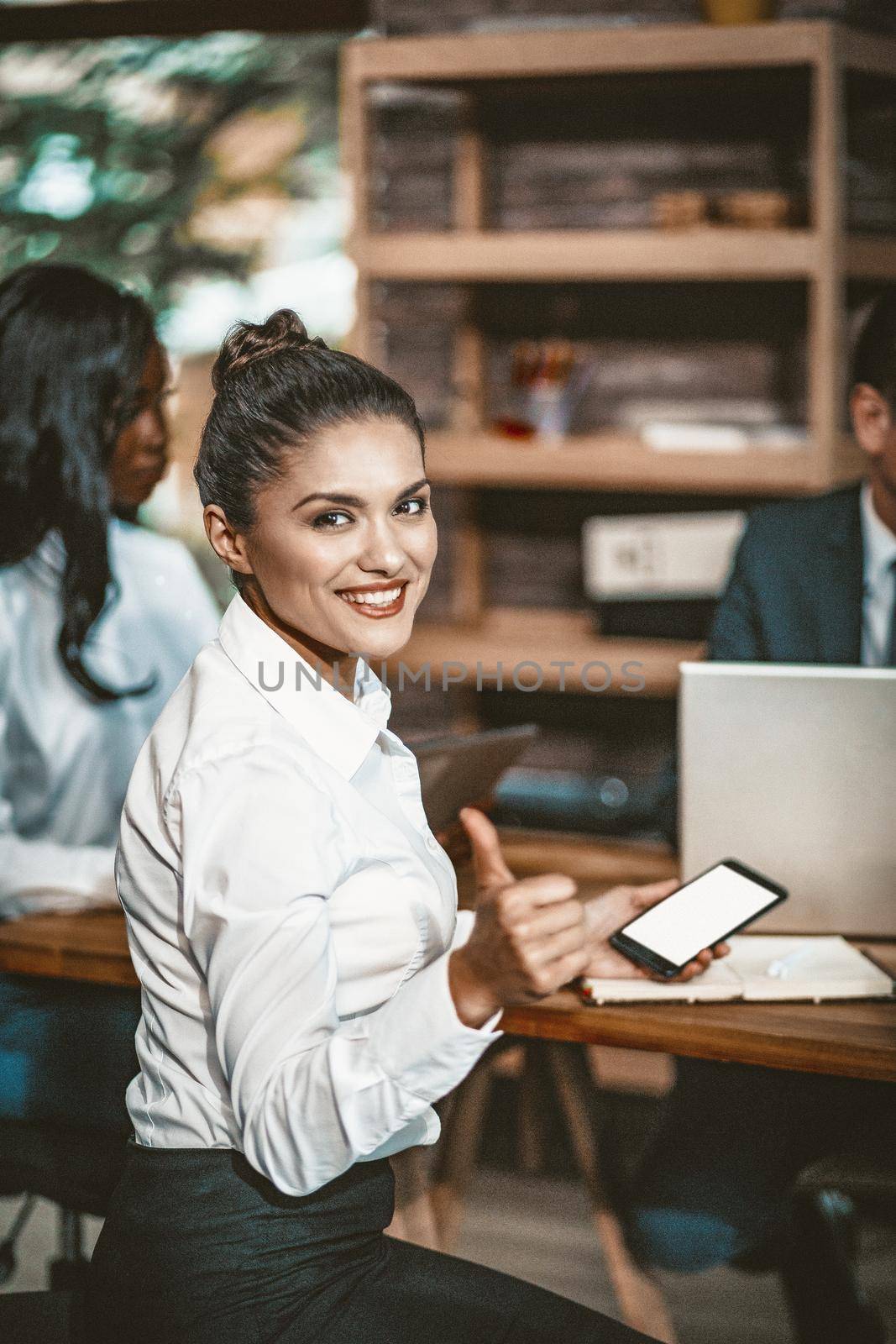 Charming Office Worker Toothy Smiles Looking At Camera, Young Business Woman In White Blouse Shows Thumb With One Hand And Holds Smart Phone With A White Screen In Other Hand, Toned Image