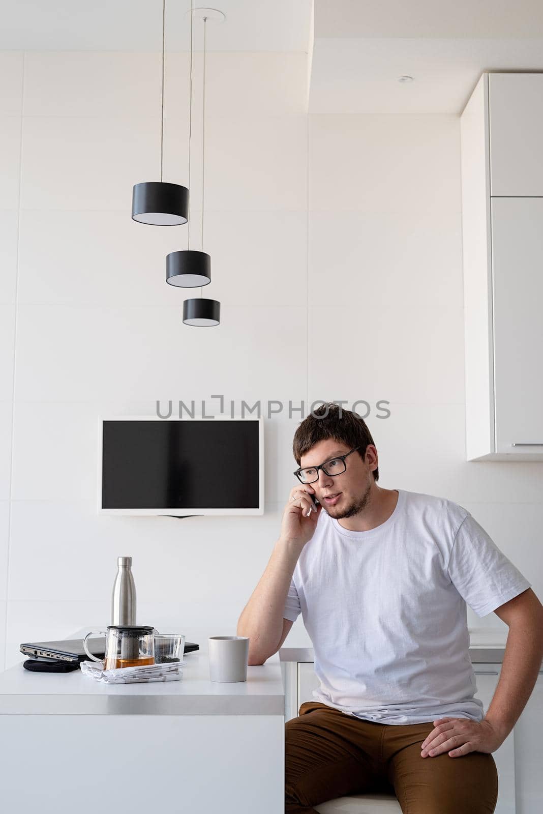 Young caucasian thoughtful man sitting at the kitchen drinking tea at home, using mobile phone