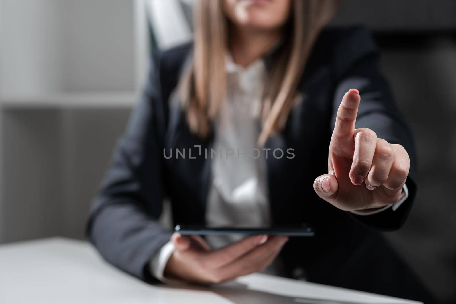 Businesswoman Holding Tablet In One Hand And Pointing With One Finger On Important Message. Sitting Woman In Office With Cellphone In Hand Presenting Informations. by nialowwa