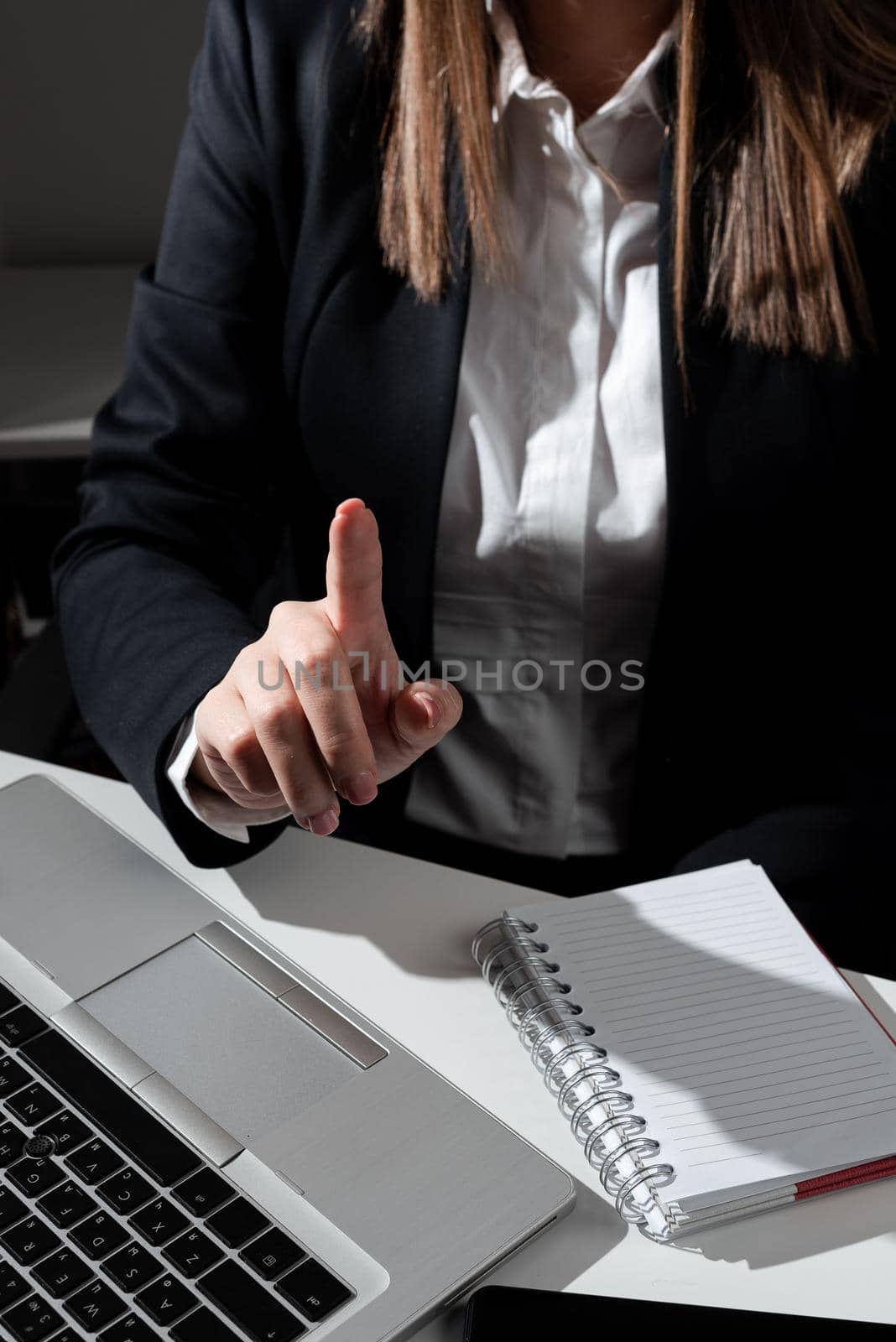 Businesswoman Pointing With One Finger On Important Message Sitting On Desk With Lap Top And Notebook. Woman In Suit Presenting Crutial Information With One Hand. by nialowwa