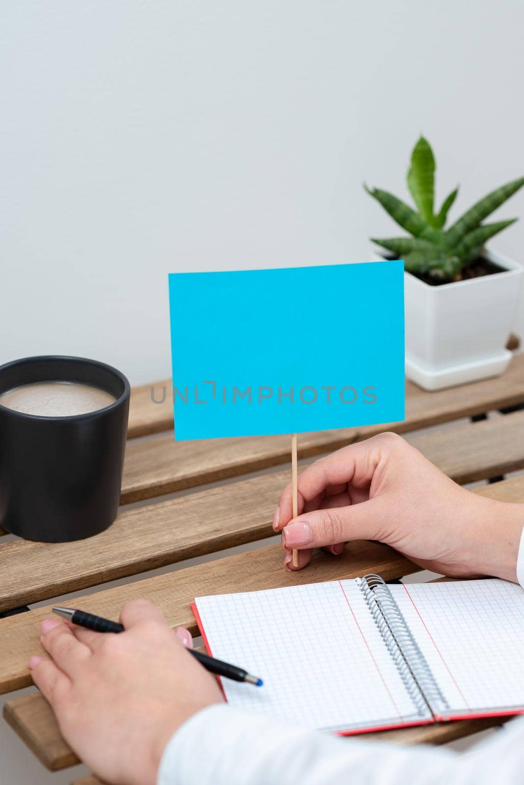 Woman Holding Pen And Important Message On Stick On Table With Notebook, Plant And Coffee. Sitting Lady With Pen In One Hand And Crutial Information In Other. by nialowwa