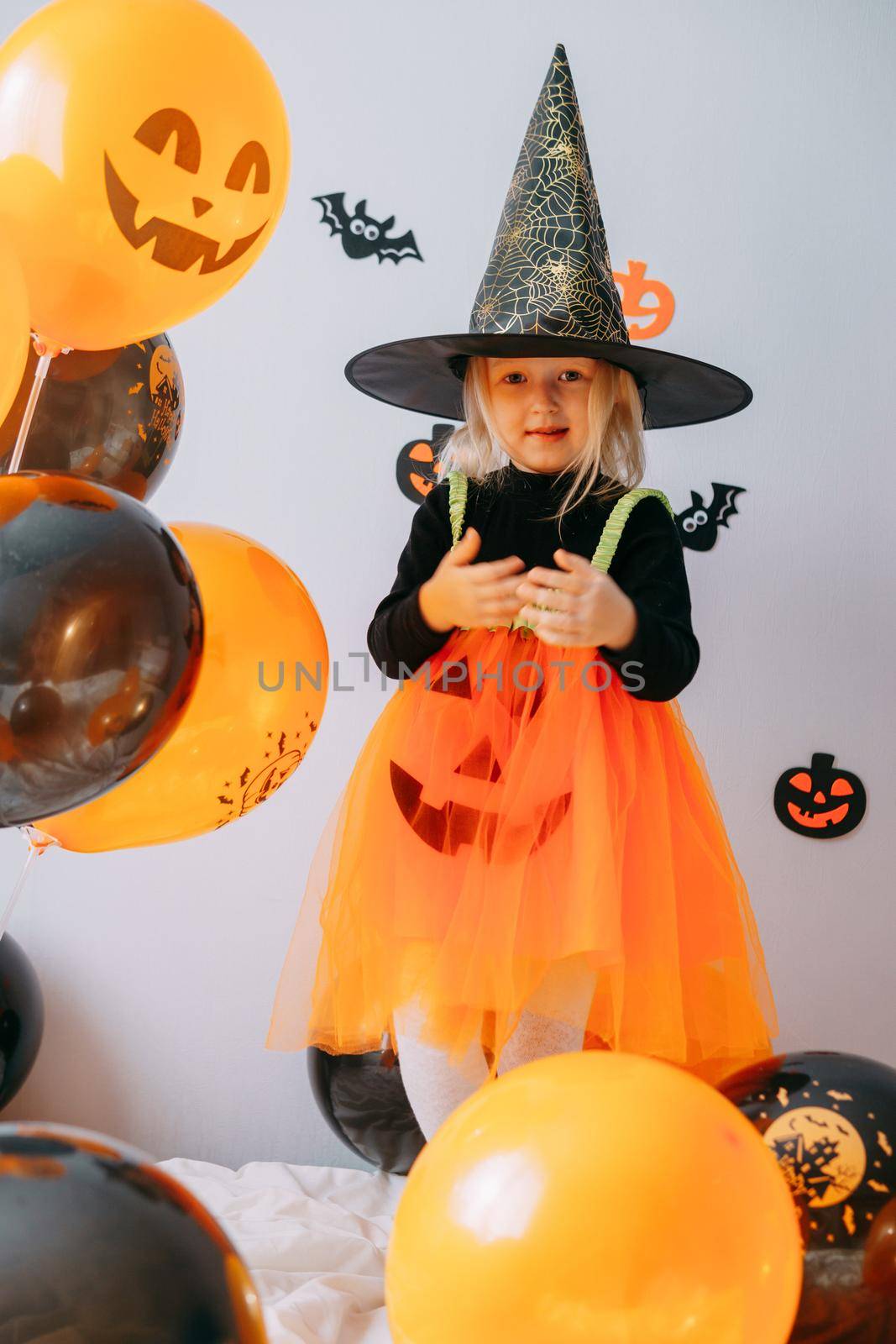 Children's Halloween - a girl in a witch hat and a carnival costume with airy orange and black balloons at home. Ready to celebrate Halloween.