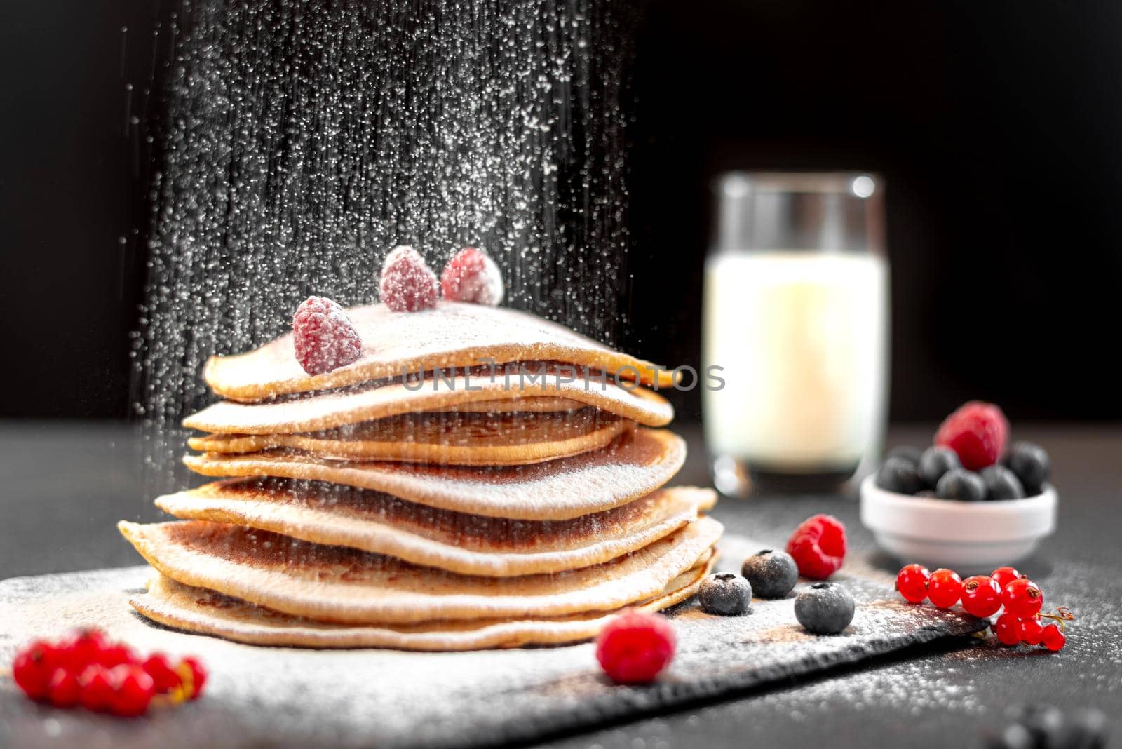 Large American pancakes on a dark background. Breakfast European. Chef sprinkles pancakes with powdered sugar on black background. Food for breakfast healthy eating. Pancakes without butter with berries. Food for vegetarians and for dieting. American Pancakes Mini. Mini pancakes with blueberries and raspberries. American breakfast on a dark background. Healthy eating for the whole family. Homemade pancakes with berries
