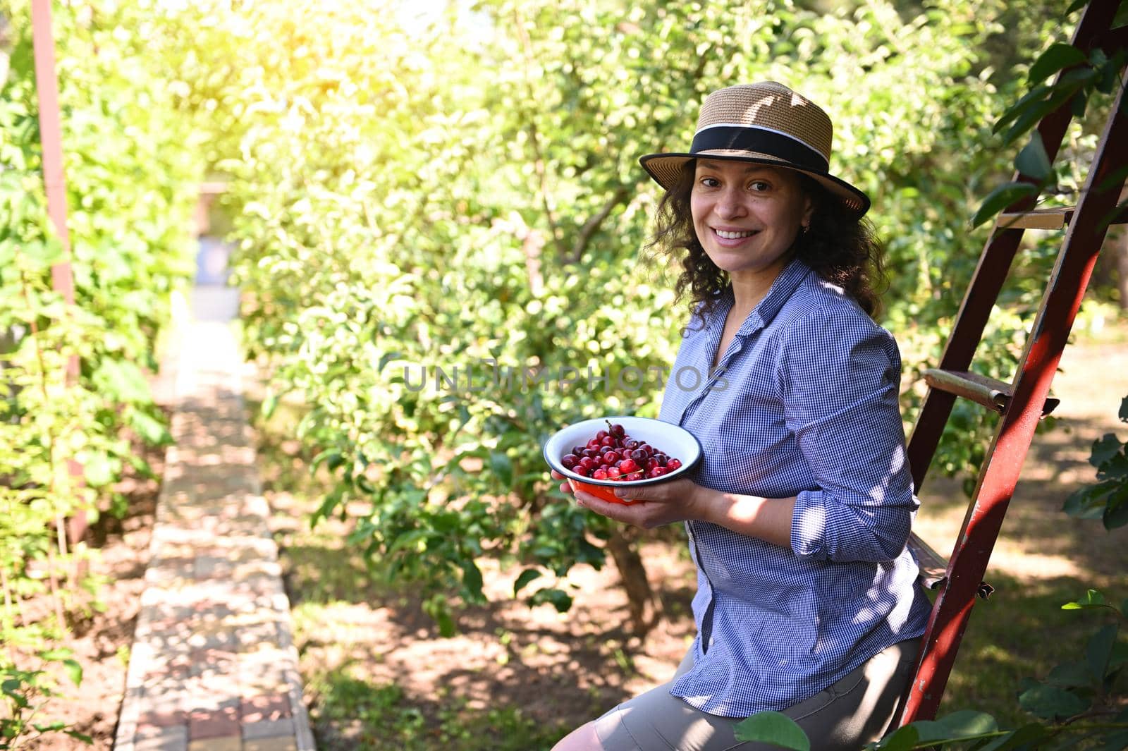 Charming woman, eco gardener smiling with a beautiful toothy smile, looking at camera, holding a bowl with freshly picked cherries in an organic orchard. Countryside summer pleasures. Eco farming