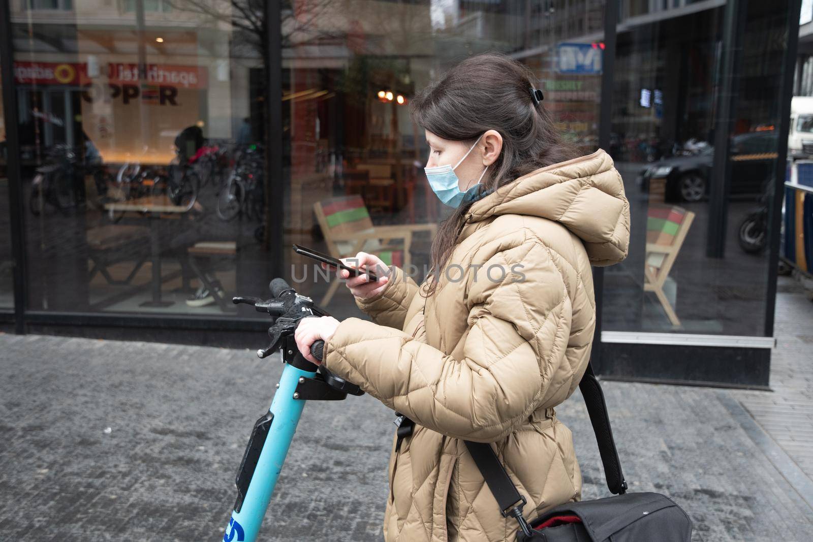 Brussels Belgium - 01.31.2022 a young woman in a warm down jacket and a mask reserves a public scooter parked near a shop on a city street. High quality photo