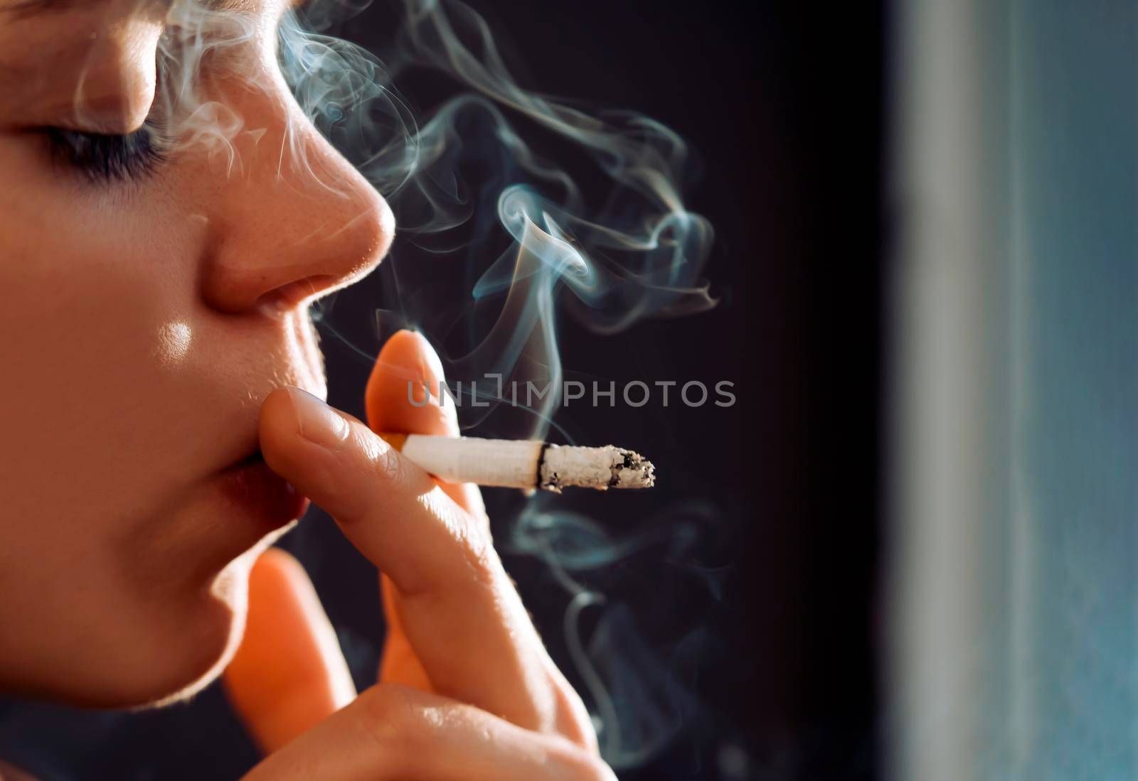 A young beauty girl is smoking, a woman is stressed and holds a cigarette in her hands, staying near the window of her apartment, close-up view.