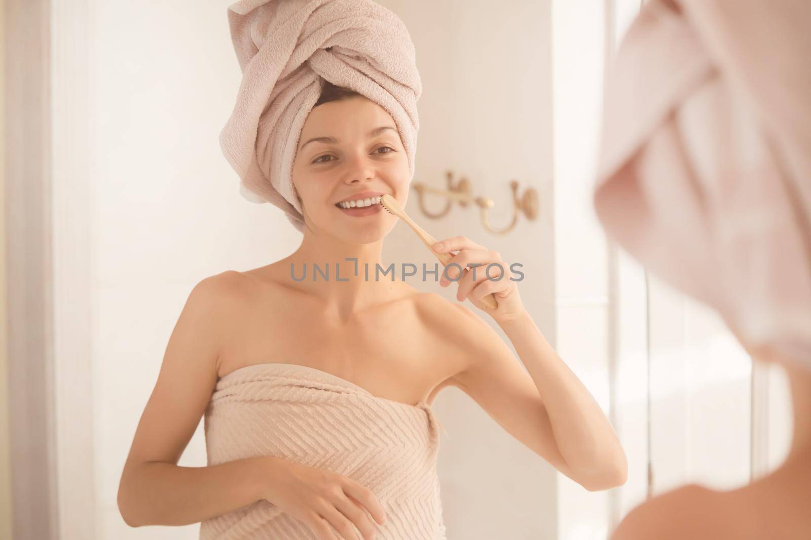 A young girl brushes her teeth near the mirror after relaxing in the bathroom, the woman is wrapped in a towel and takes care of the health and beauty of her body and teeth, gums.