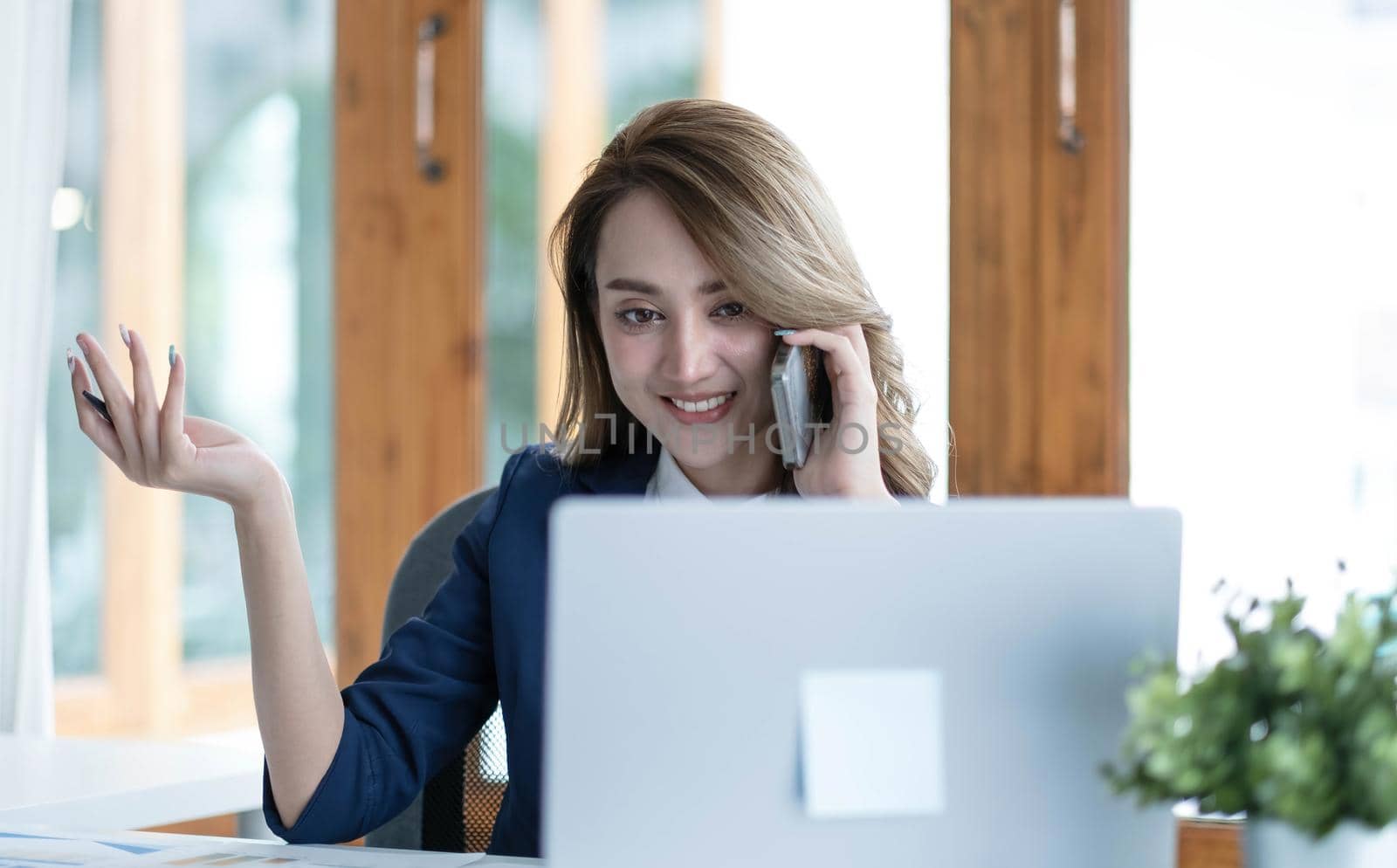 Happy young Asian business woman sitting smiling and talking on mobile phone in office..