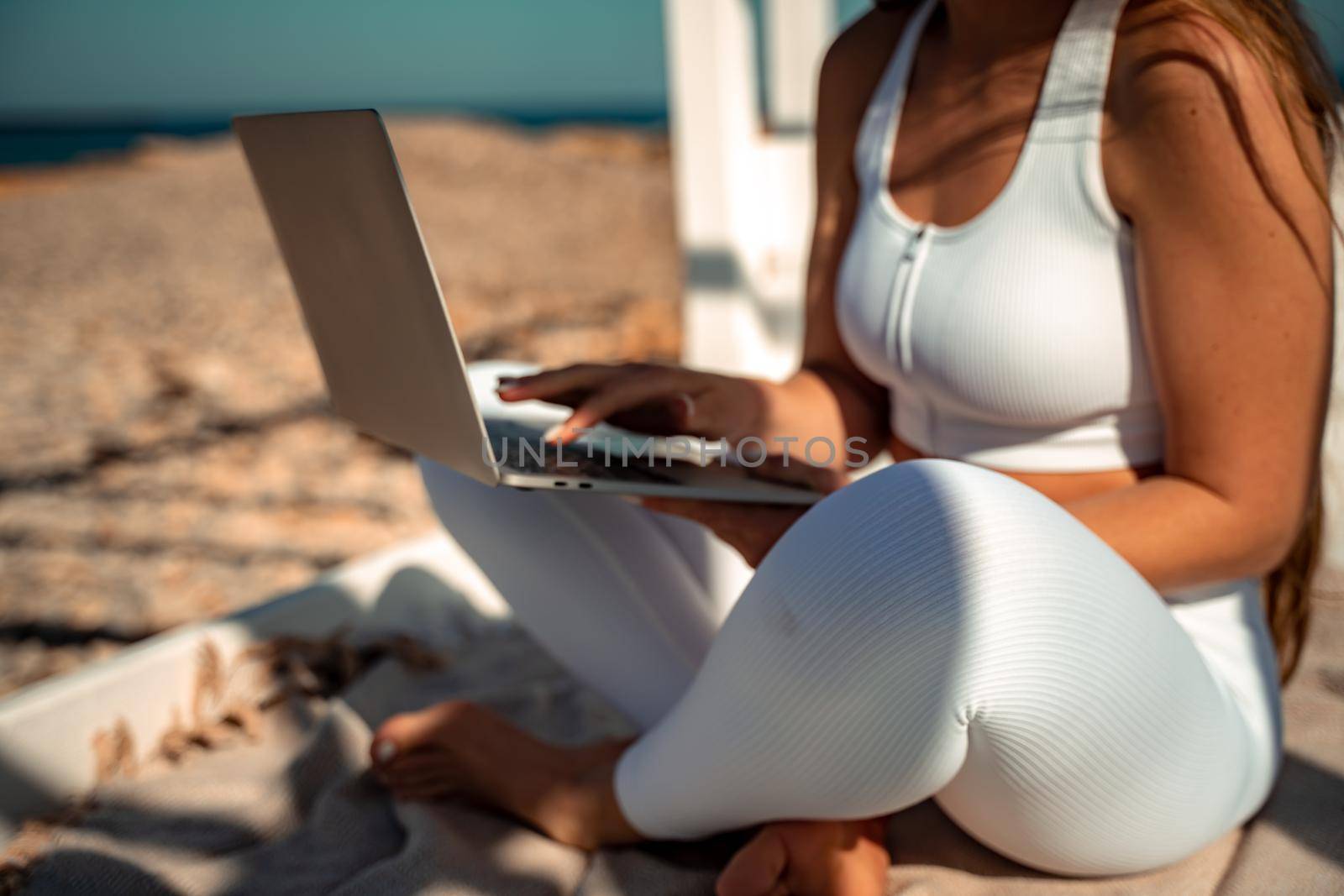 young beautiful woman with dark hair in white overalls sits on the sand near the sea and uses a laptop,