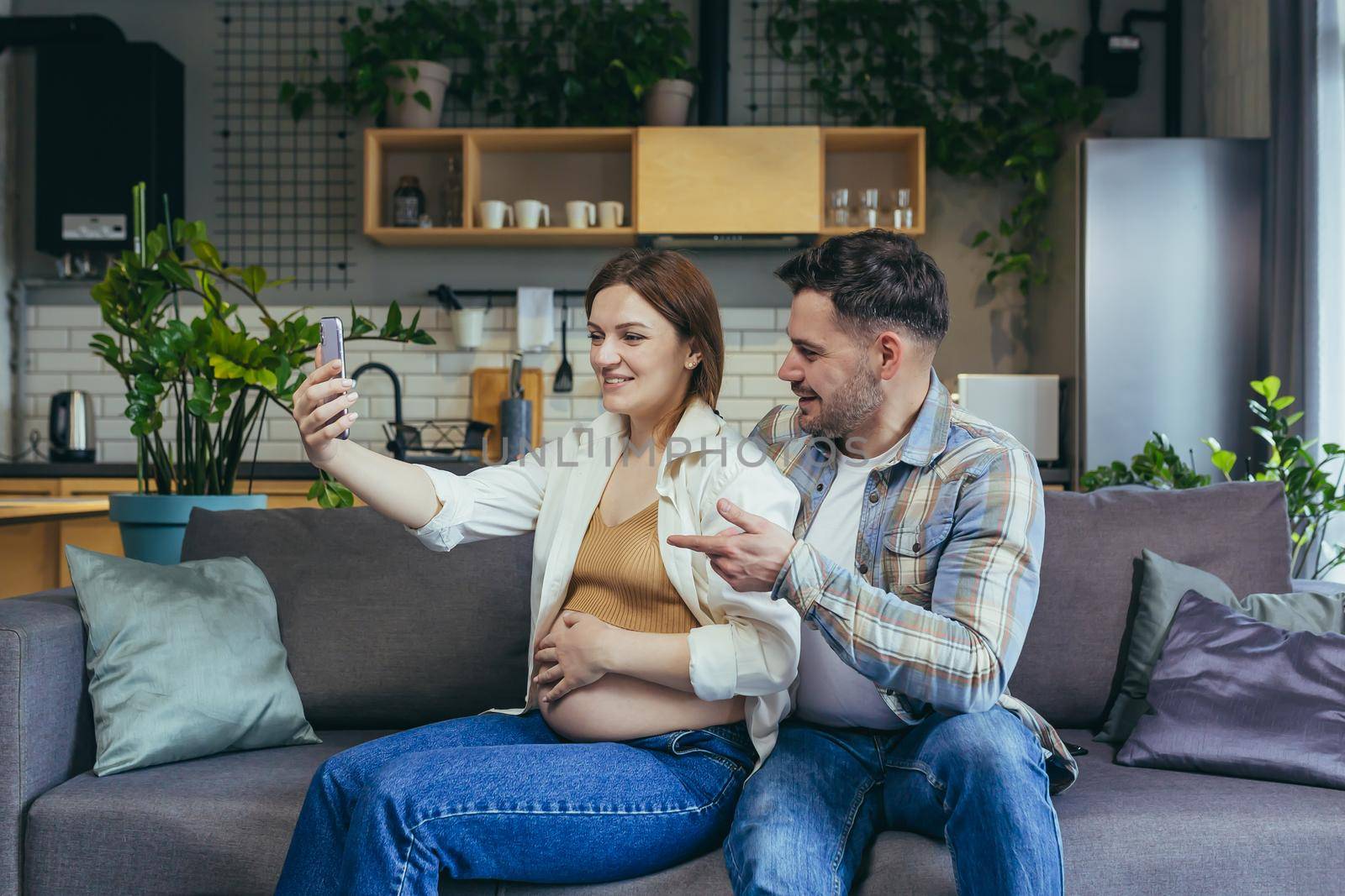 Happy young married couple man and pregnant woman using phone for video call, hugging and sitting on sofa smiling and looking at smartphone screen