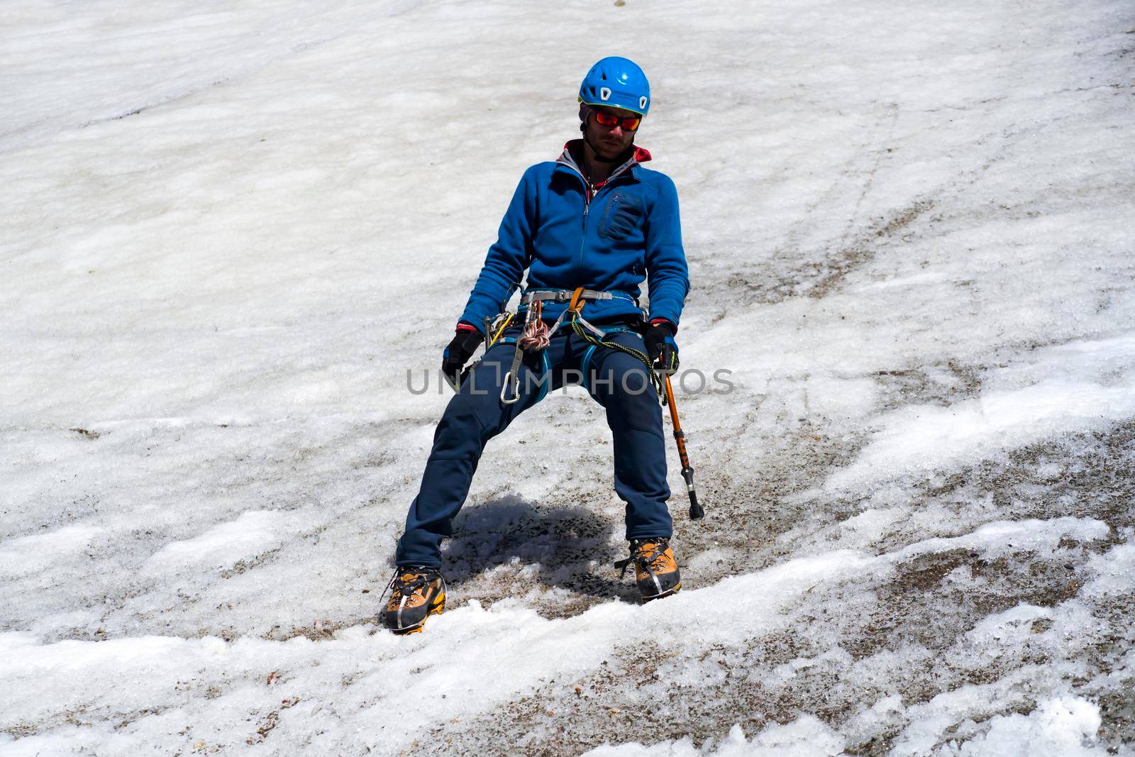 Young stylish man in sunglasses, a traveler with ice tools while hiking in the mountains, a tourist with climbing equipment, in a helmet, crampons walks on a glacier, a snow-covered mountain.