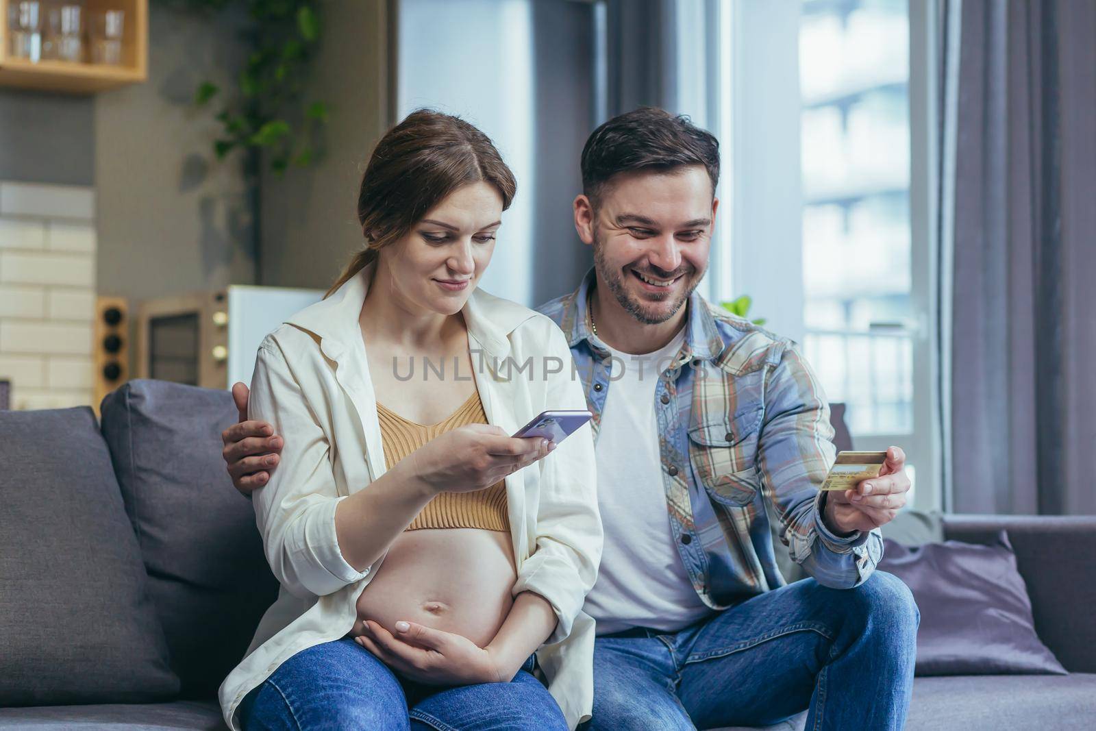Young married couple husband and pregnant woman sitting together on the couch and hugging using the phone for online shopping in the online store, holding a bank credit card