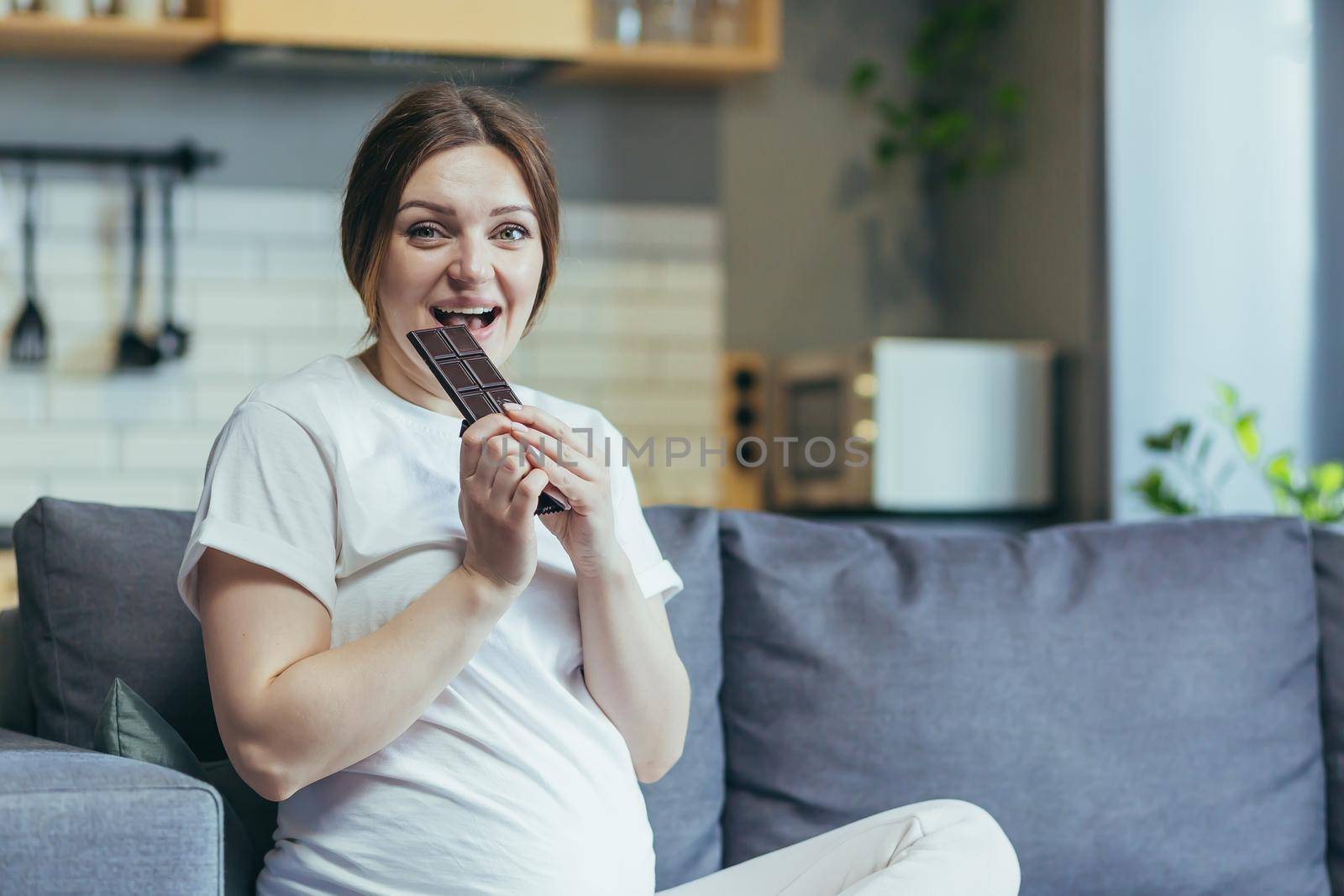 Young pregnant woman at home sitting on the couch eating chocolate