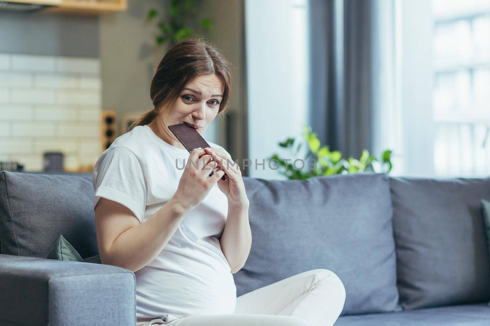Young pregnant woman at home sitting on the couch eating chocolate