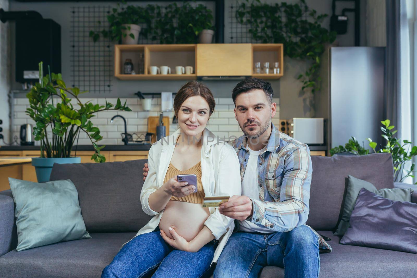 Young married couple husband and pregnant woman sitting together on the couch and hugging using the phone for online shopping in the online store, holding a bank credit card