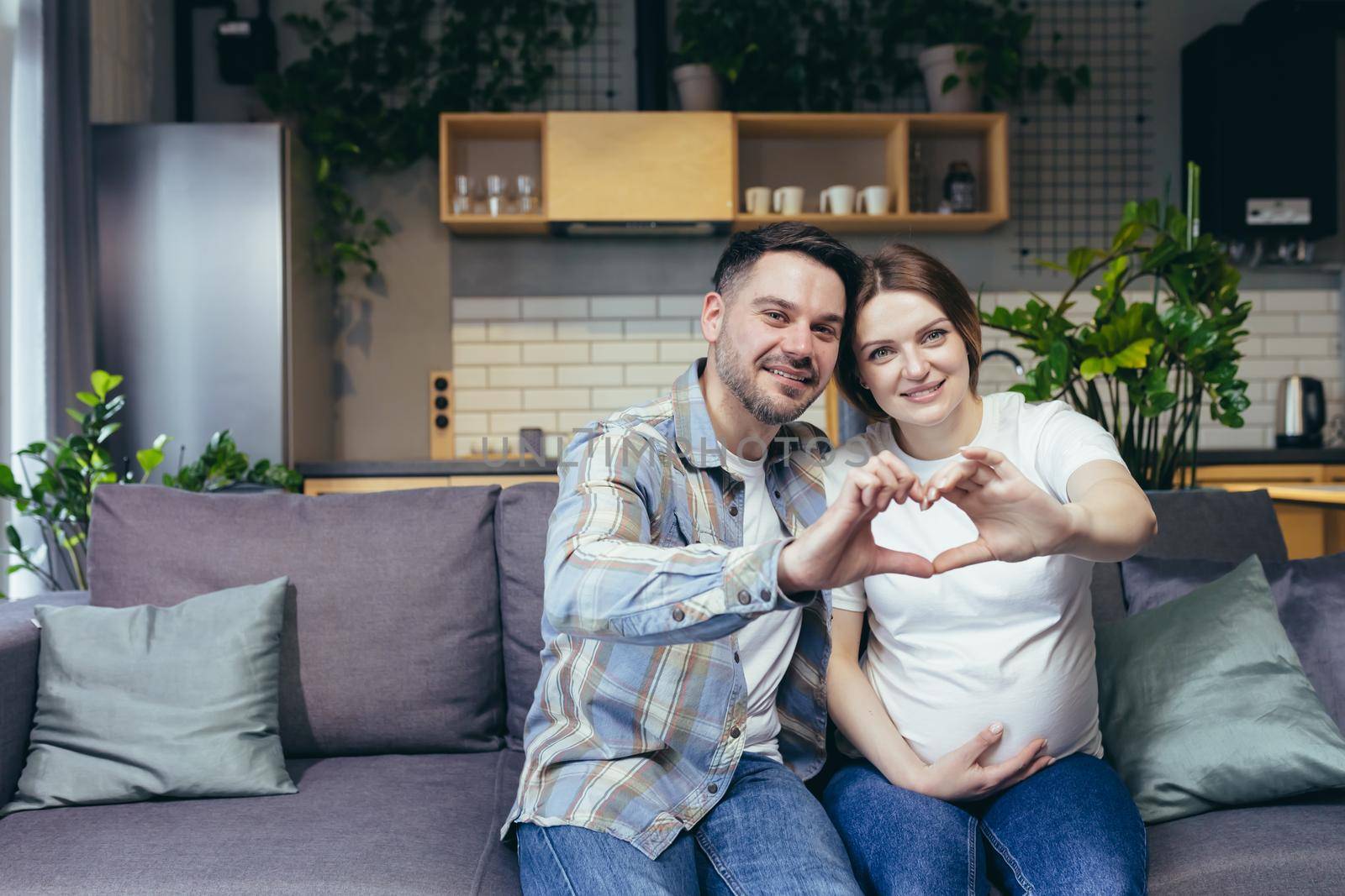 Portrait of a young happy family. Husband and pregnant woman waiting for the birth of a child sitting on the couch at home, hugging, making hands with heart, looking at the camera, smiling