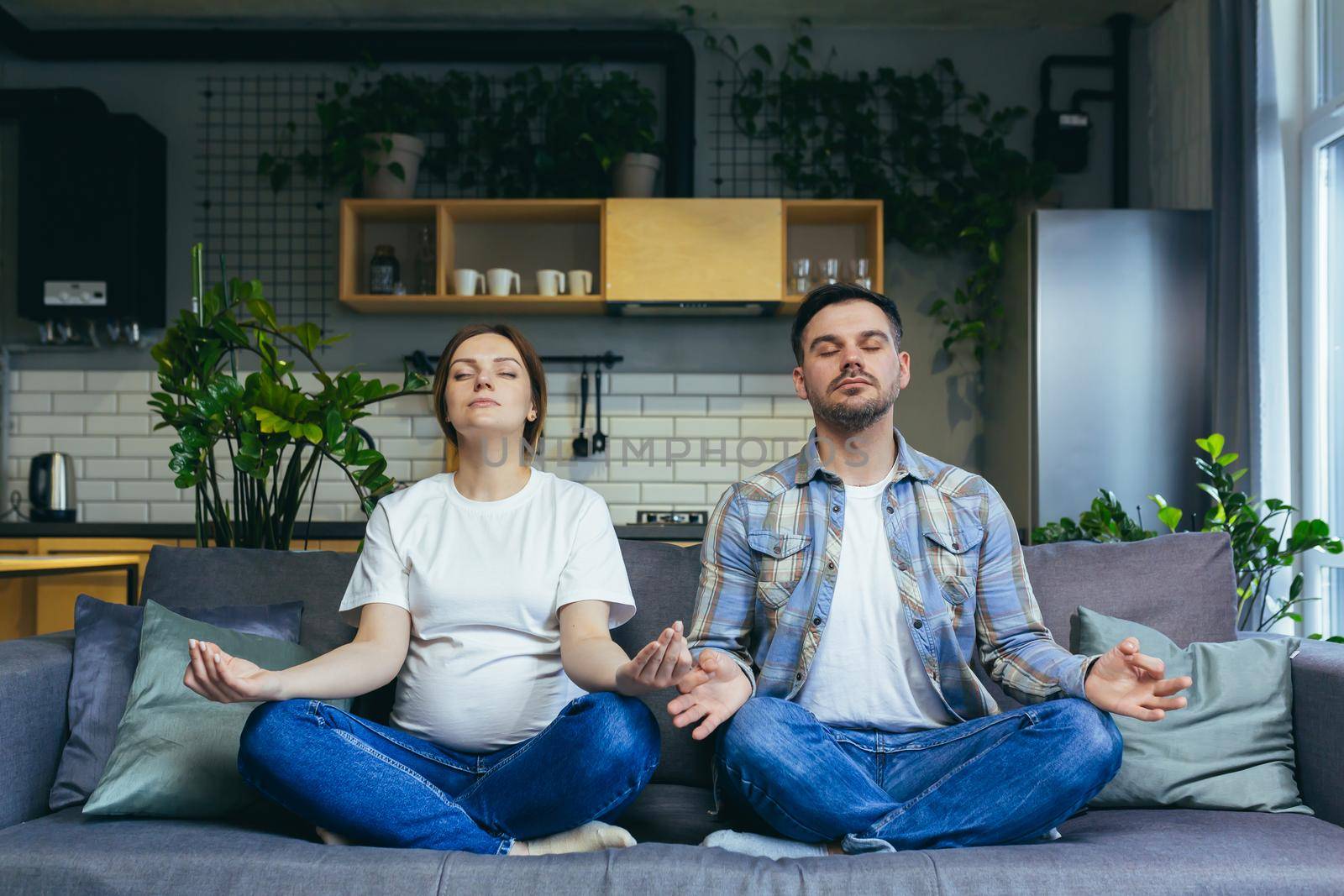 A pregnant man and woman are meditating in a lotus position on the couch