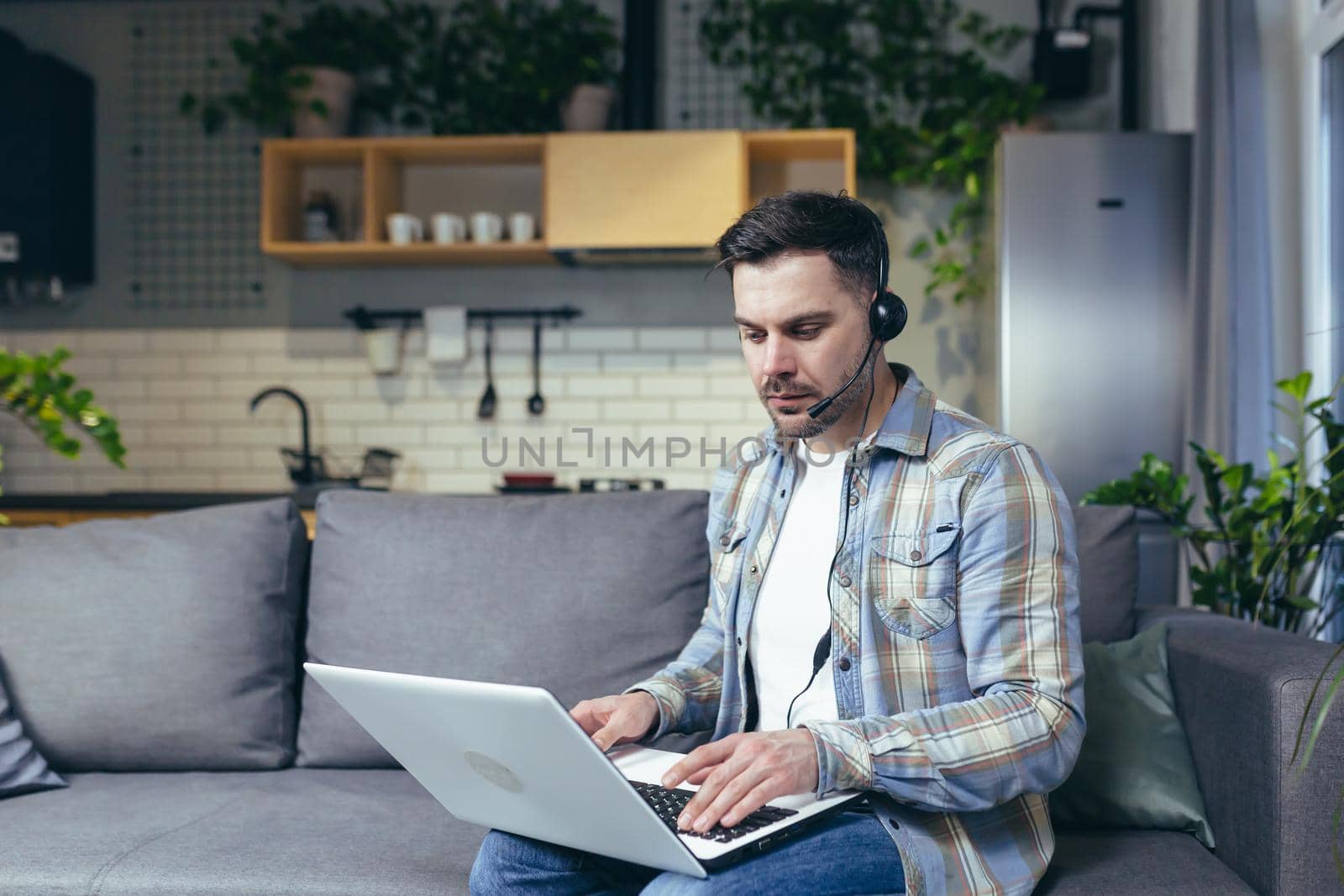 Young man working from home with laptop and in headphones. Talks on video call. Sitting on the couch.