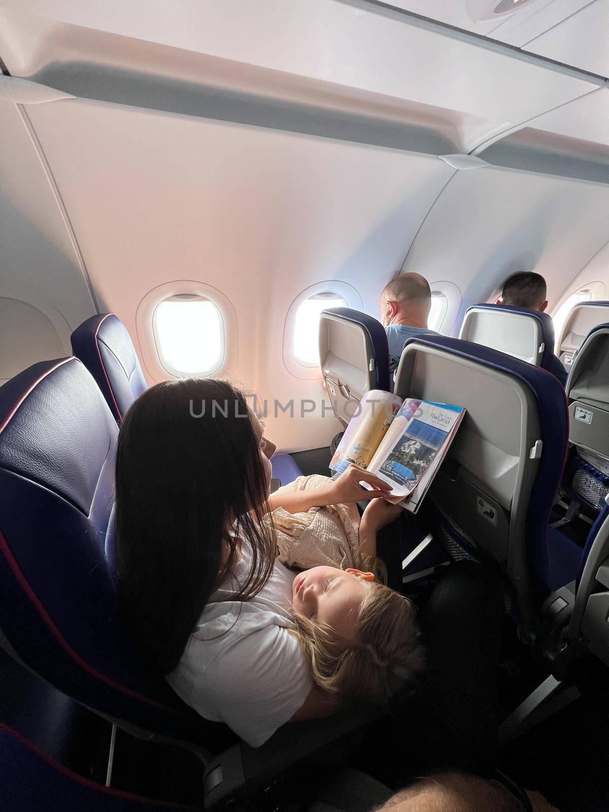 Mom with a sleeping daughter in her arms sits in an airplane seat and reads a magazine by Nadtochiy