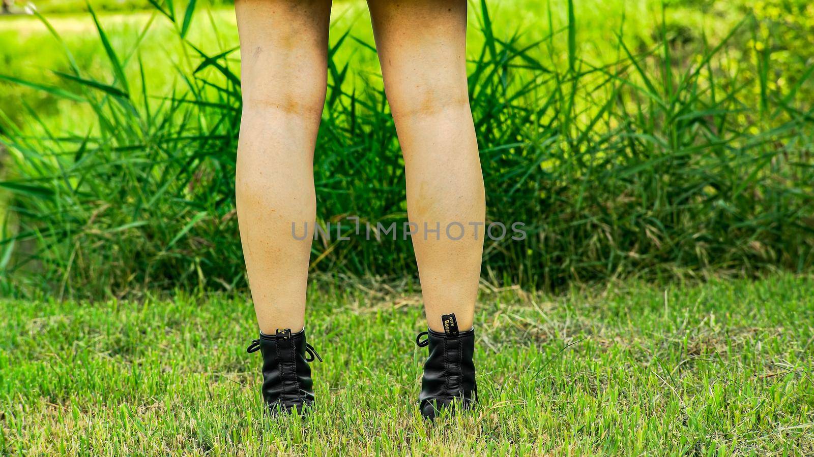 Girls bare legs back view with black long boots walk slowly in the green meadows