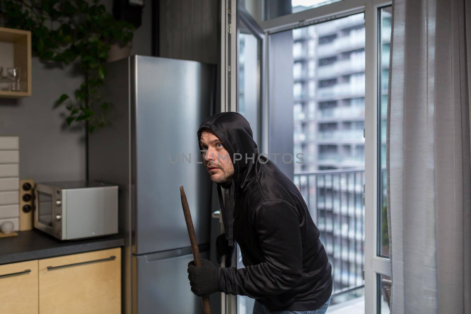 Young male robber in dark clothes carefully and quietly enters through the balcony window into the apartment, house. Holds scrap metal, fittings.