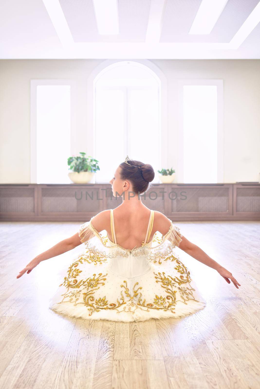 Back view of beautiful young ballet dancer in cream dress sitting and warms up her hands on wooden floor in ballet studio