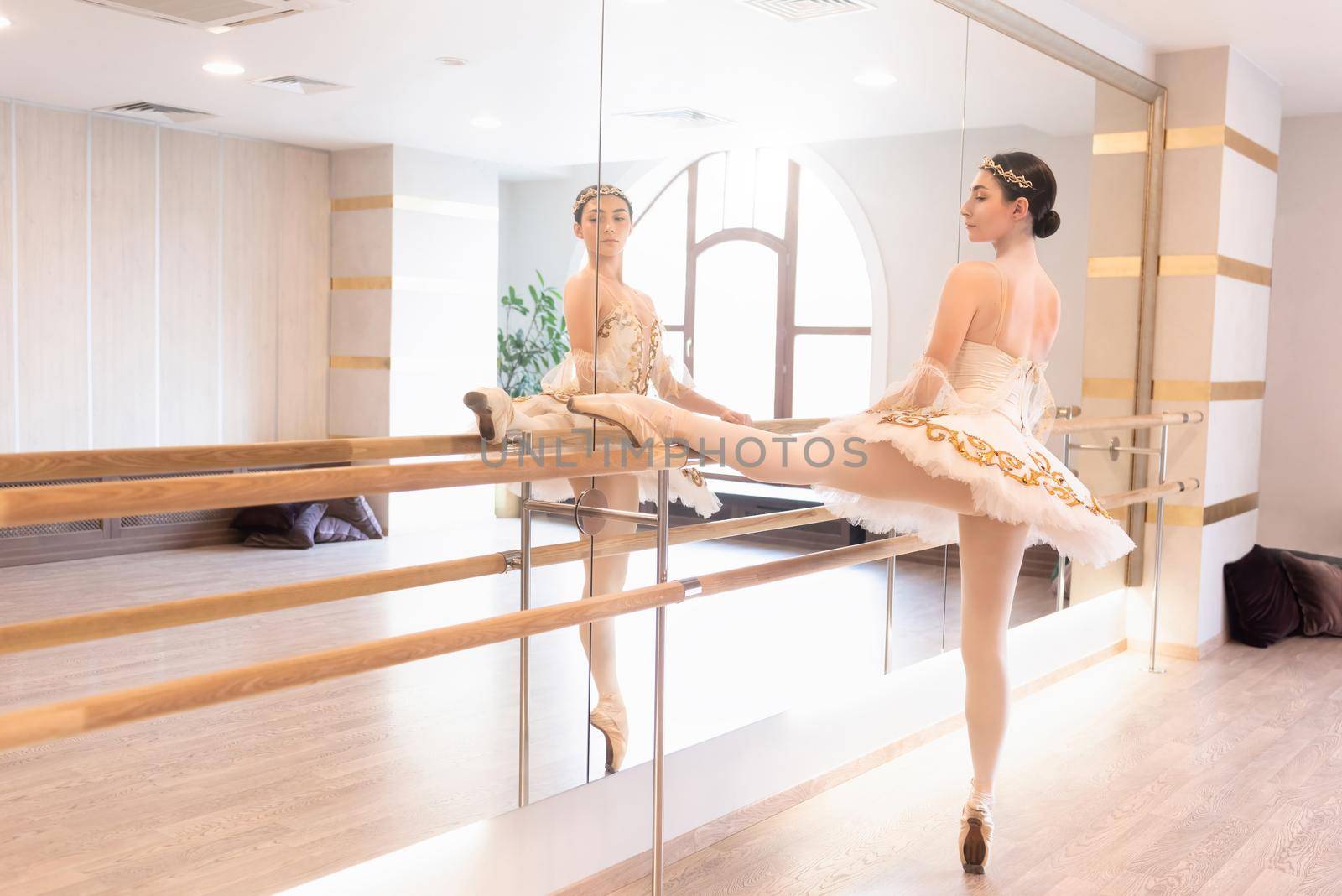 full length view of ballerina in cream dress and pointe shoes training at barre near mirror by Nickstock