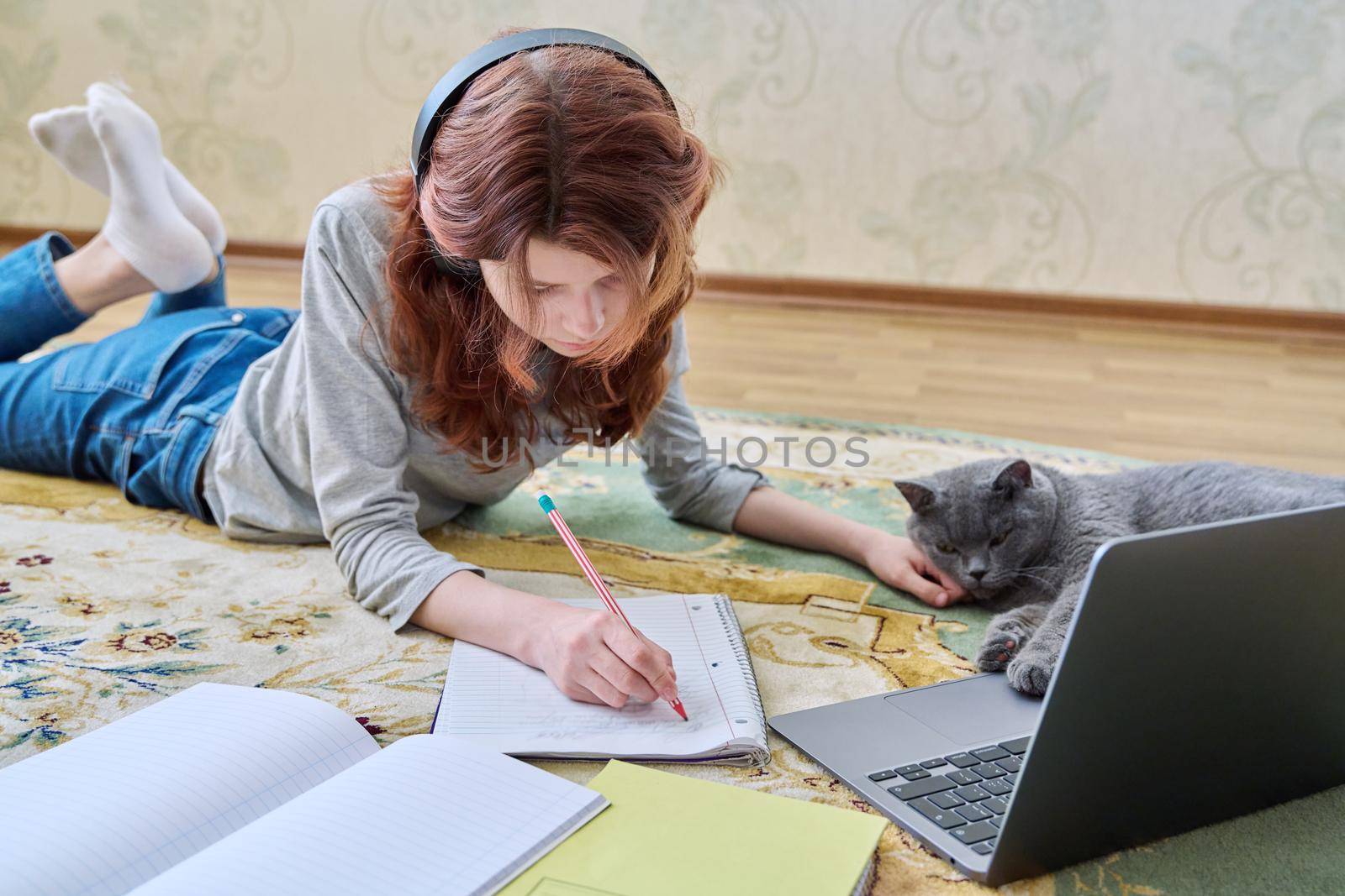 Preteen girl studying in headphones with laptop lying on floor with cat. Young female student using laptop, writing in notebook. School, learning, distance education, homework, pets lifestyle concept