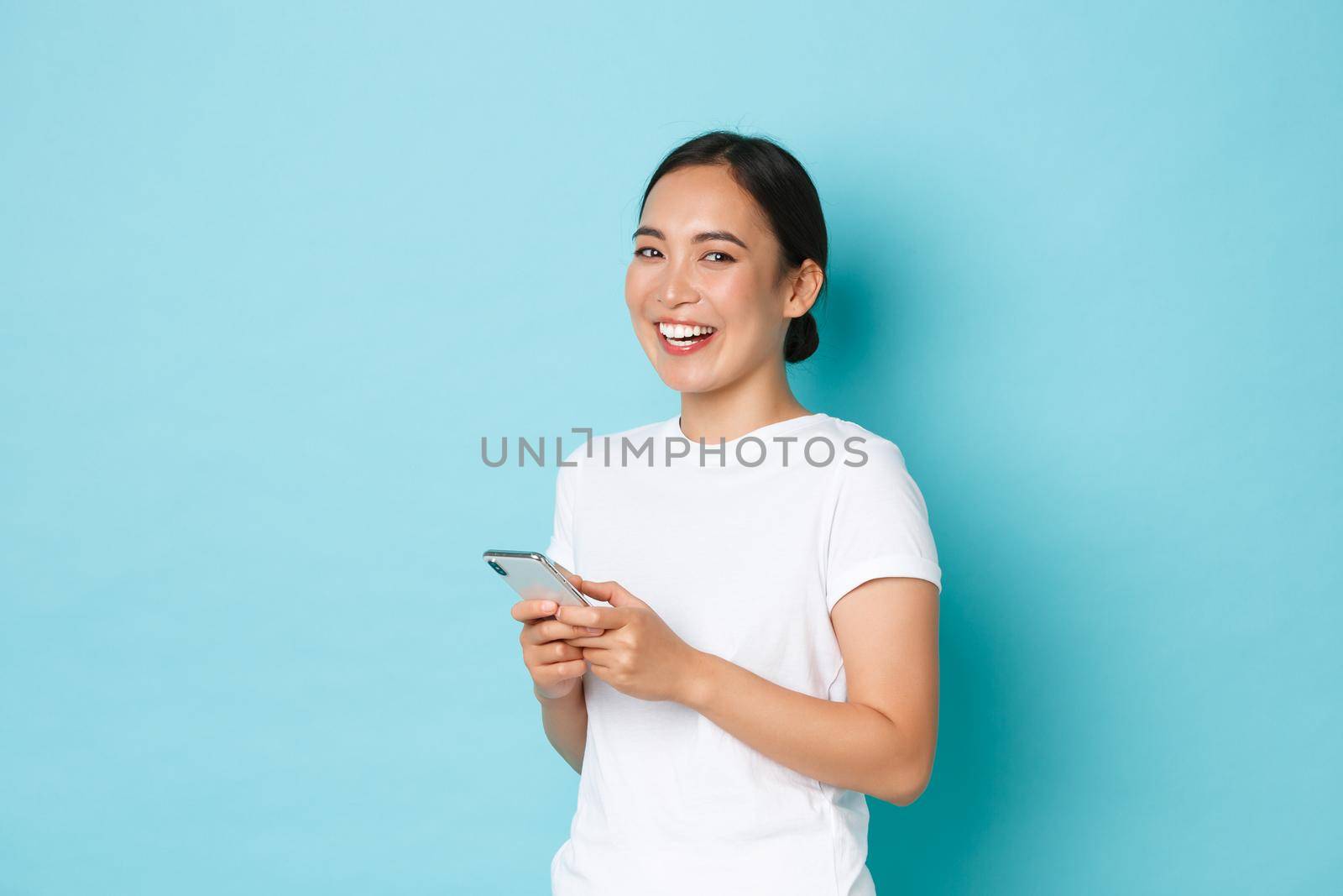 Lifestyle, technology and e-commerce concept. Side view of attractive asian girl using mobile phone, texting, messaging or chatting with friends online, starting to laugh and look at camera.