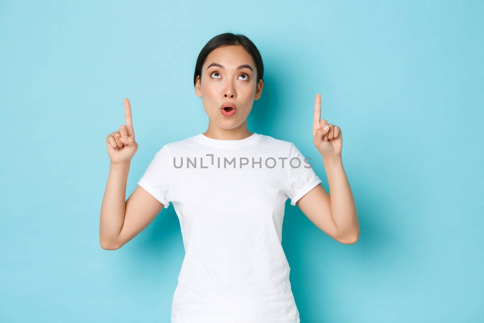 Surprised and impressed asian girl in white t-shirt looking and pointing fingers up at something interesting and amazing. Beautiful korean girl 20s found awesome promo offer, over blue background by Benzoix