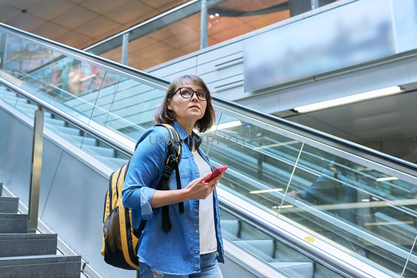 Middle-aged woman with backpack smartphone in her hands walking up stairs, near escalator in modern station building. Urban architecture, urban lifestyle, people concept