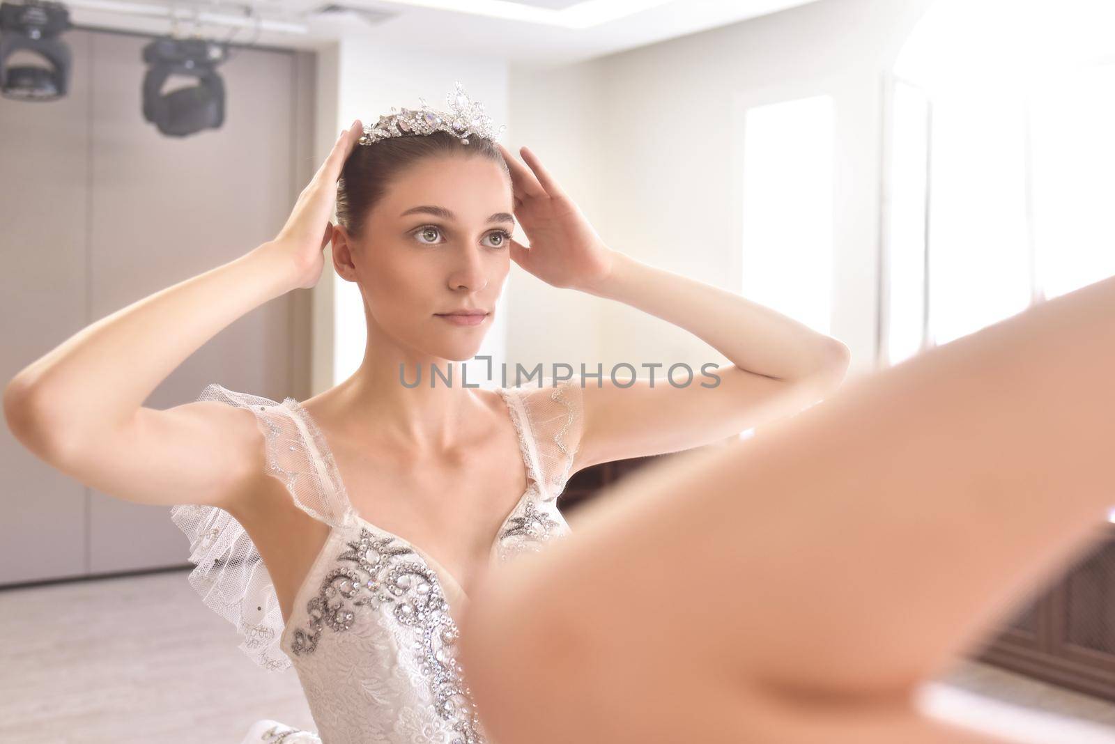 A young woman in a cream dress applies makeup and wears a crown in front of the mirror. The concept of beauty