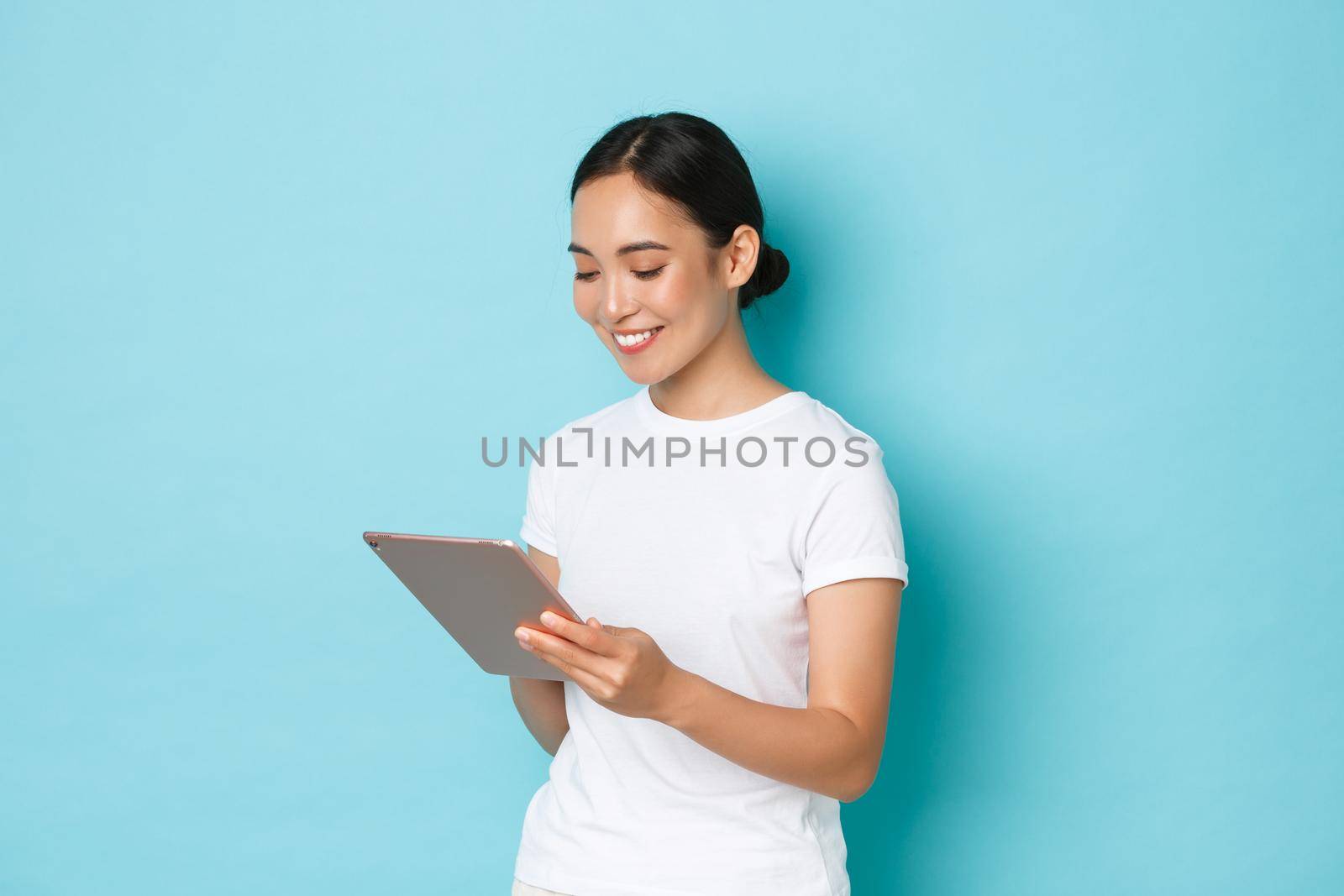 Portrait of smiling beautiful asian girl, female 3d artist or freelancer using digital tablet and looking pleased at device screen, shopping online or browsing over light blue background.