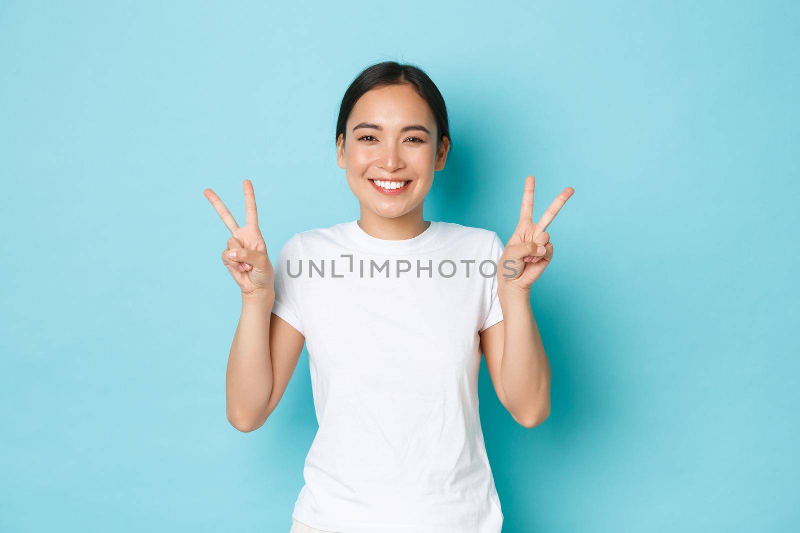 Beauty, fashion and lifestyle concept. Portrait of beautiful asian girl in white t-shirt standing over blue background, smiling and showing kawaii peace gesture, sending positivity and joy by Benzoix