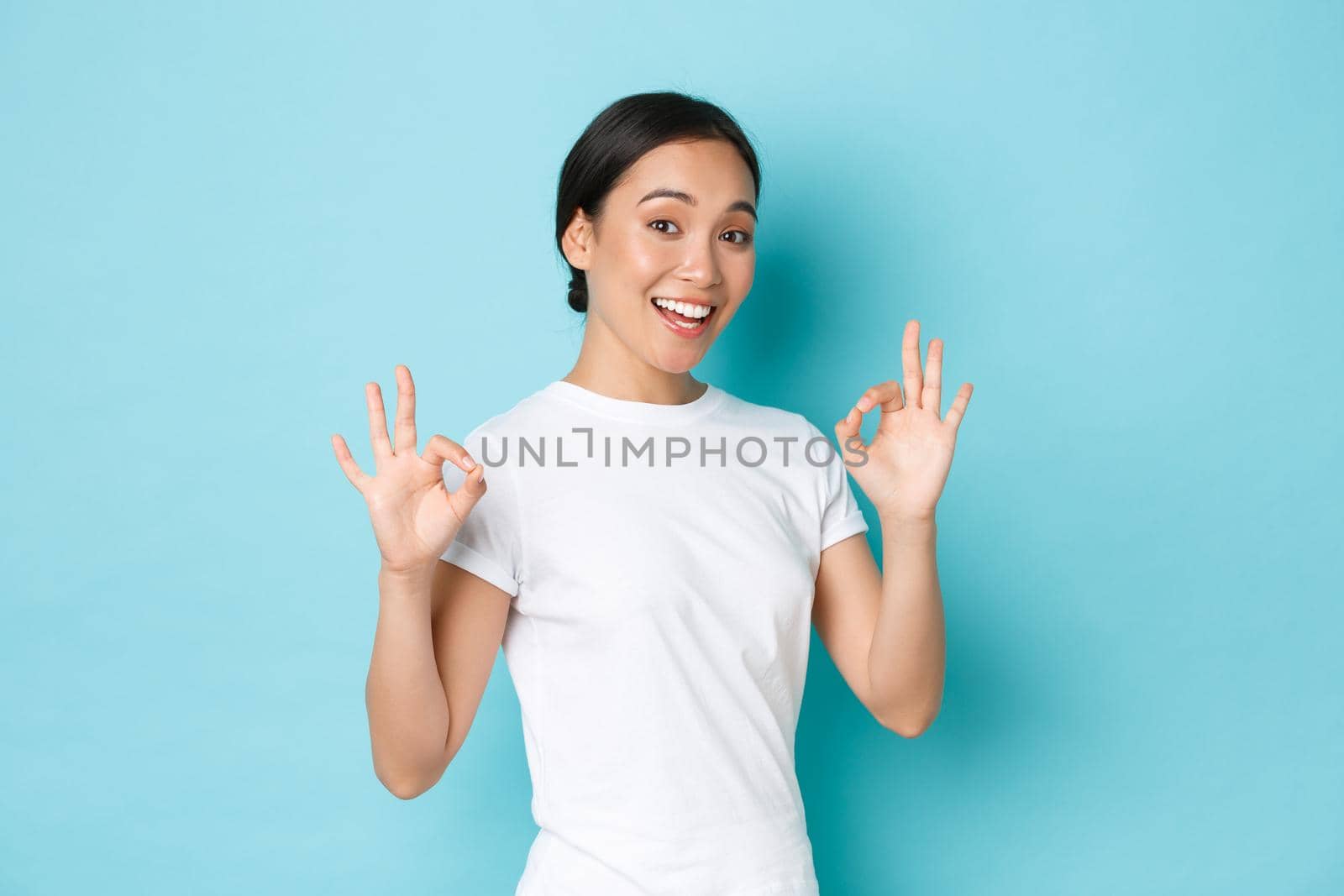 Pretty confident asian smiling girl ensure everything OK, under control. Woman encourage all good, showing okay gesture and nod in approval, guarantee quality, standing satisfied blue background.
