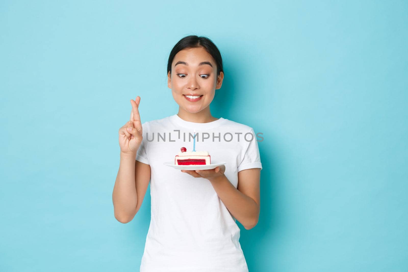 Holidays, lifestyle and celebration concept. Hopeful pretty asian girl in white t-shirt making wish for her birthday, looking thoughtful and cross fingers for good luck while holding b-day cake.