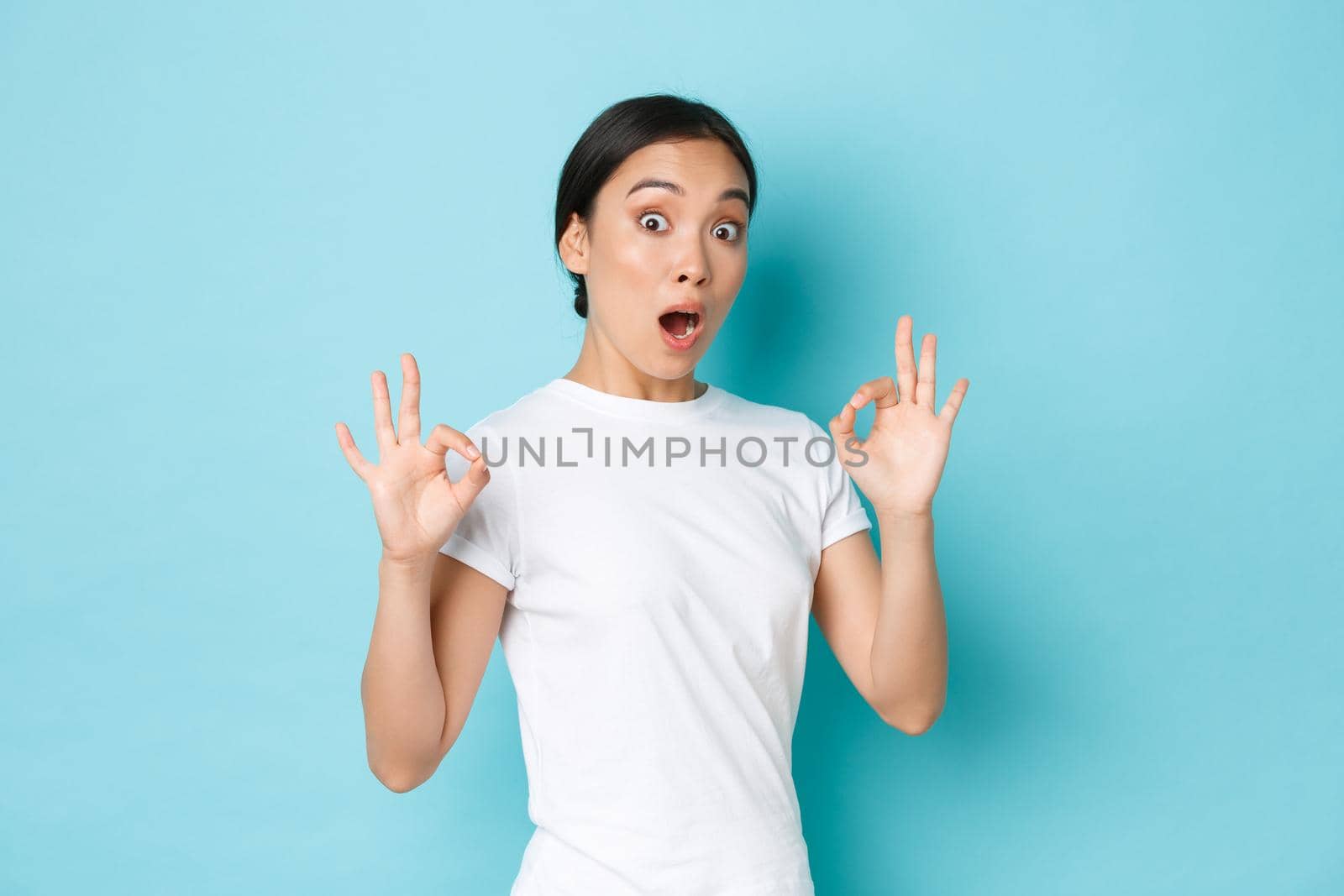 Excited and impressed asian girl in white t-shirt look surprised and astonished with amazing, perfect service, showing okay gesture and looked astounded over blue background by Benzoix