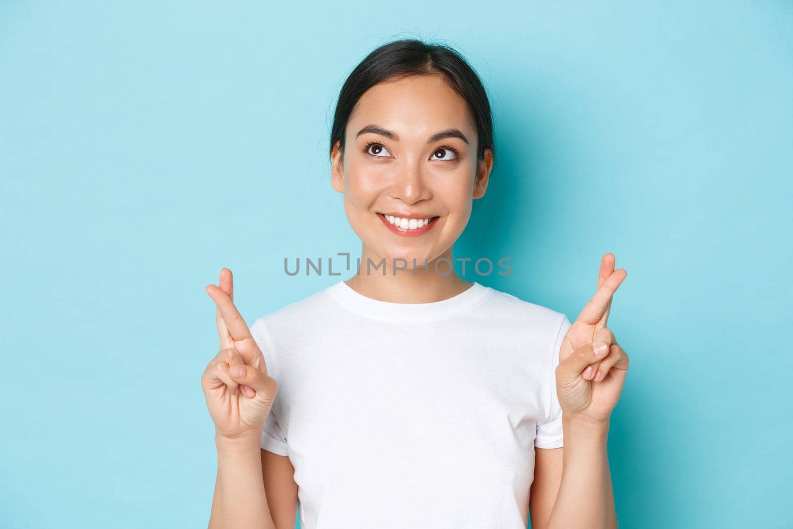 Close-up of hopeful dreamy asian girl making wish, looking upper left corner thoughtful, standing light blue background with fingers crossed for good luck, praying or anticipating positive news.